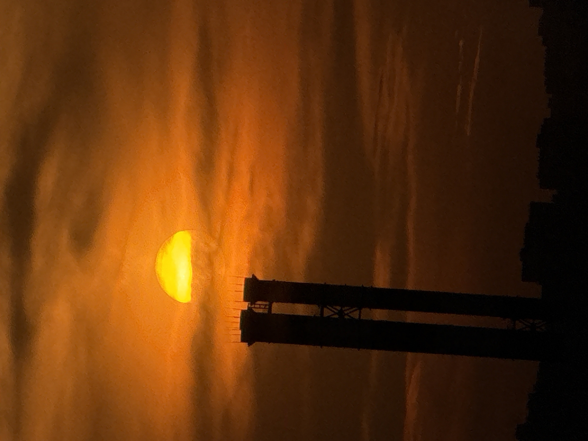 The image captures a sunset or sunrise over an urban landscape in Moscow, Russia. The sky is dominated by intense orange and red hues, indicative of a low sun angle, with visible horizontal cloud formations partially obscuring the upper atmosphere. A bright, semi-circular disk of the sun, glowing yellow-orange, is visible through the clouds in the upper-central portion of the frame, appearing to rest just above a prominent silhouetted industrial structure. This structure consists of two tall, dark, cylindrical stacks, identifiable as the CHPP-25 (ТЭЦ-25) or Zapadnaya (Western) Thermal Power Plant, located in the Ochakovo-Matveevskoye district of Moscow. The two stacks are connected by a visible cross-braced framework and a vertical ladder positioned between them. Multiple slender vertical protrusions, likely lightning rods or antennae, are present at the top of each stack. The lower portion of the image reveals a dark, silhouetted urban skyline composed of buildings and trees. No human figures or discernible actions are present in the scene.