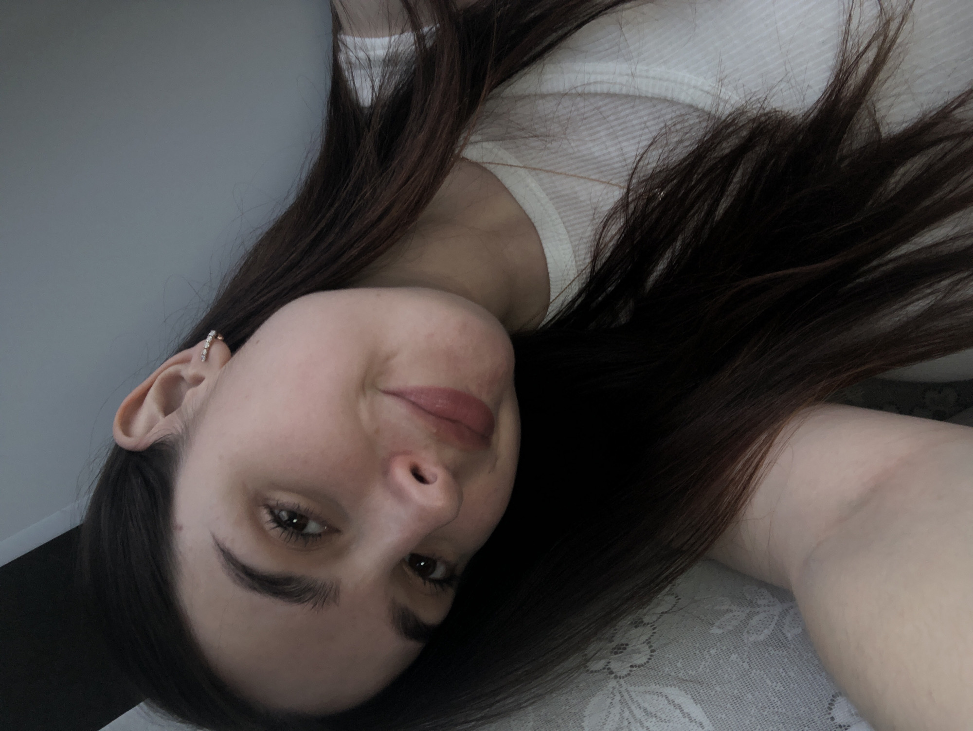 A woman with long, dark brown hair is lying down, facing the camera. She has fair skin, dark eyebrows, and brown eyes. She is wearing a white ribbed tank top and a small earring in her left ear. The background appears to be a light-colored wall and a patterned surface, possibly a bedspread. The image is slightly rotated. There is no evidence to suggest this image was taken in Noviy, Russia.
