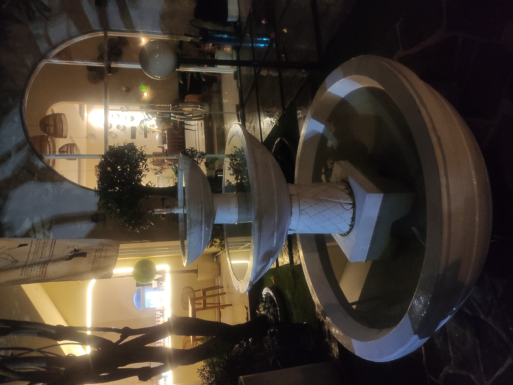 The image shows a three-tiered water fountain located in what appears to be an outdoor dining area in East Jakarta, Indonesia. The fountain is constructed of light-colored stone or concrete and has a geometric base. Each tier holds water, and small plants are placed around the fountain.
In the background, a building is visible, reflecting the interior through a curved glass window. The interior is well-lit and shows a cafe or restaurant setting with tables, chairs, and hanging light fixtures. There are no visible people in the immediate vicinity of the fountain. The ground around the fountain is paved with dark stone. Tree branches are present in the foreground, partially obscuring the view. The scene is lit by both indoor and outdoor lighting.