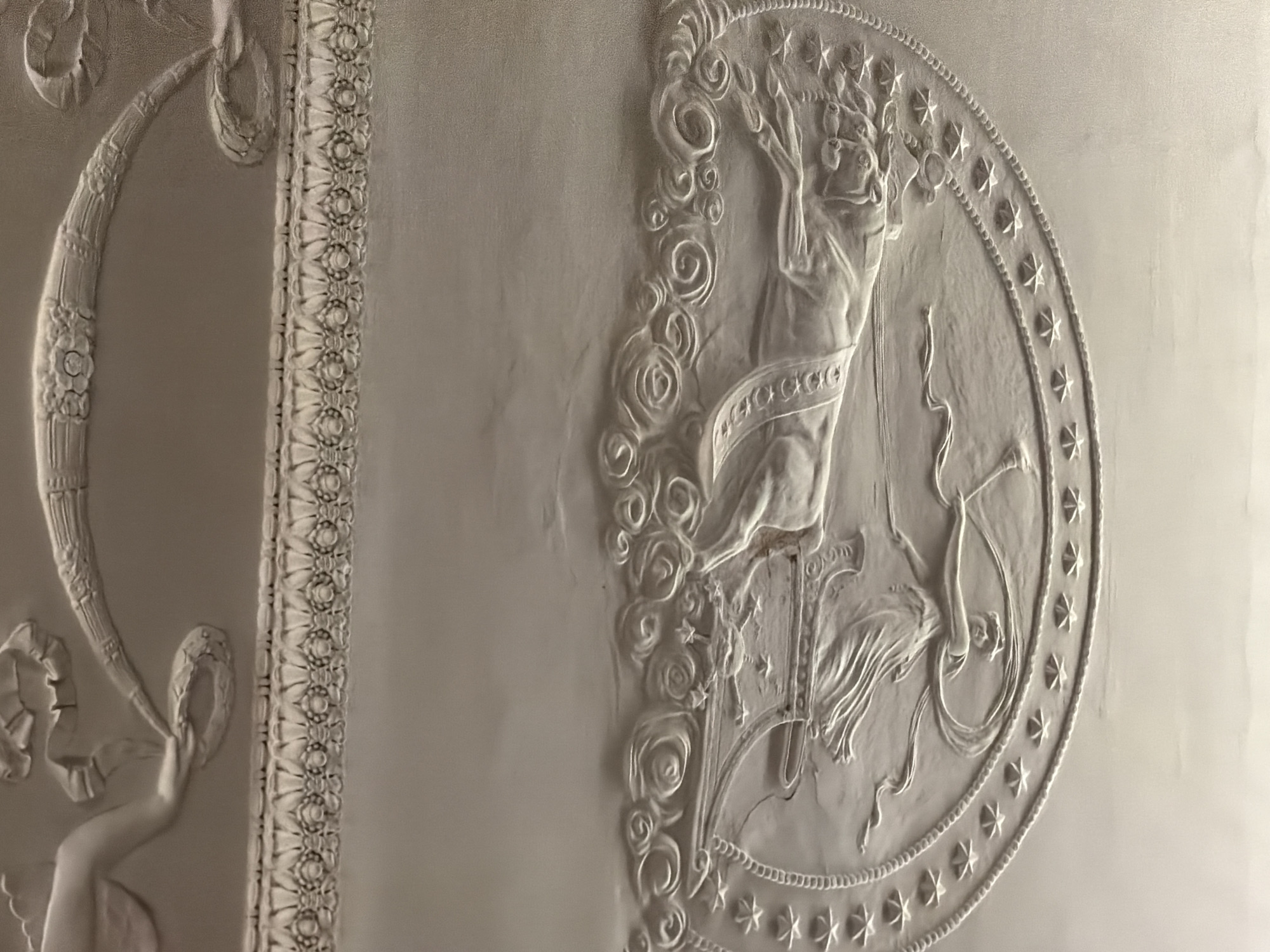 The image displays intricate white plasterwork in relief, likely adorning a ceiling or wall, indicative of classical or Neoclassical interior architecture.
The central subject is a relief sculpture of a classical female figure, possibly a goddess or allegorical representation, positioned within a large circular frame. She appears to be standing or dancing, with flowing drapery and an elaborate crown or headdress adorned with stars. Her arms are raised, and she seems to be holding a stylized object, possibly a lyre or a branch. To her right, a bow and quiver are depicted, suggesting a connection to figures like Diana or Artemis, goddesses of the hunt. The circular frame around her is bordered by a continuous line of small, multi-pointed stars and a beaded edge. Outside this frame, to the left of the figure, there are swirling, rose-like floral patterns that transition into a vertical decorative panel.
This vertical panel, running down the left side of the main image, features a repetitive pattern of intertwined floral motifs and geometric shapes, creating a highly ornate border. Further to the left, another large, curved decorative element is visible, featuring a thick, twisted vine-like form from which ribbons and smaller floral or leaf motifs emanate. At the bottom of this curved element, a smaller, kneeling figure (possibly a putto or cherub) is subtly integrated into the design.
The entire scene is rendered in a monochromatic palette of white and light grey, emphasizing the three-dimensional depth and texture of the plasterwork. The craftsmanship suggests a historical and opulent setting, consistent with grand buildings found in locations like Saint Petersburg. There is no visible text in the image.