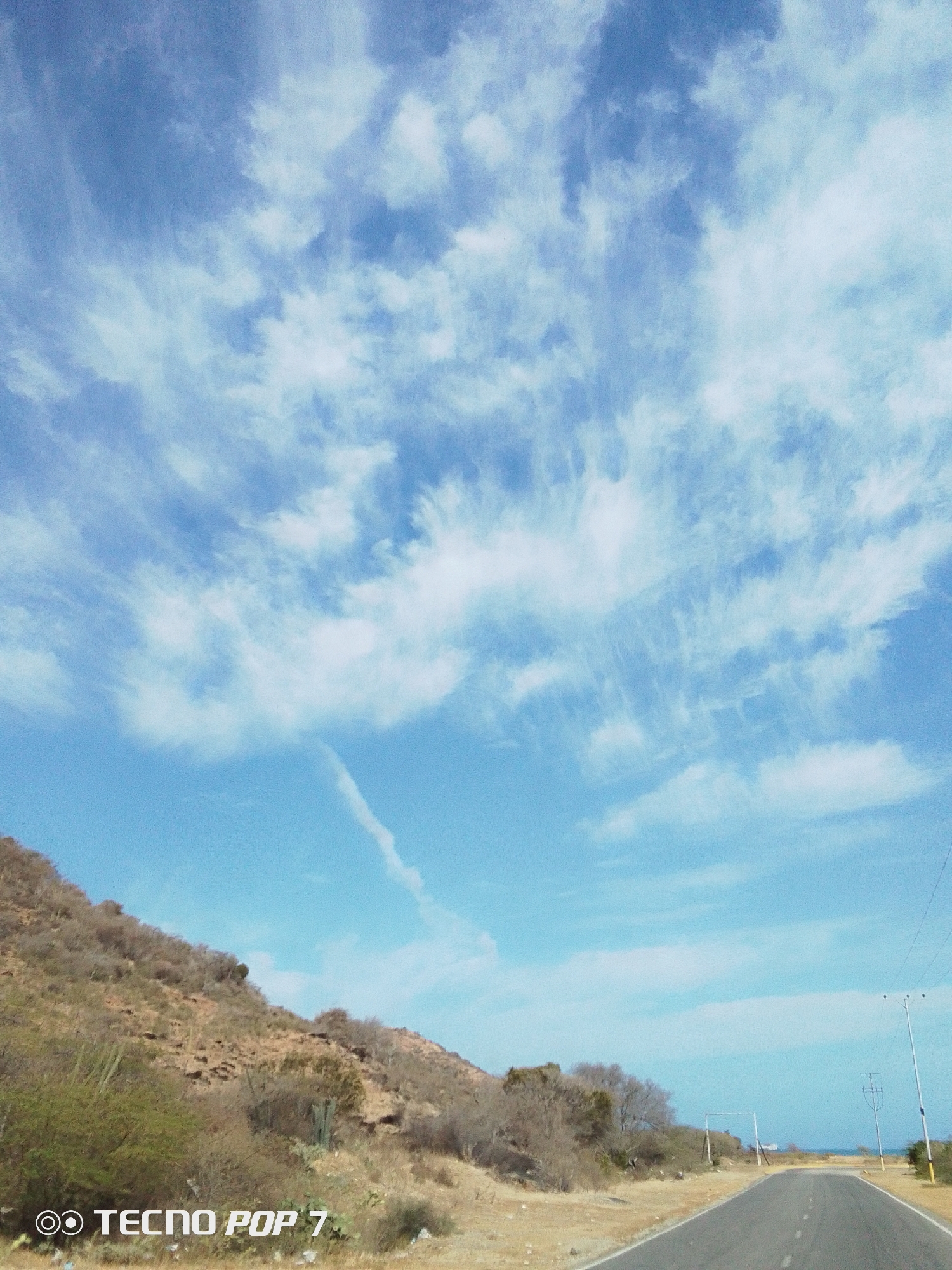 The image depicts a road leading towards the sea under a cloudy sky. Along the left side of the road is a dry, sloping hillside sparsely covered with vegetation. Utility poles are visible on the right side of the road in the distance. The image includes the location "Pedro González, Venezuela" in its implicit information. The TECNO POP 7 watermark is visible at the bottom left corner.