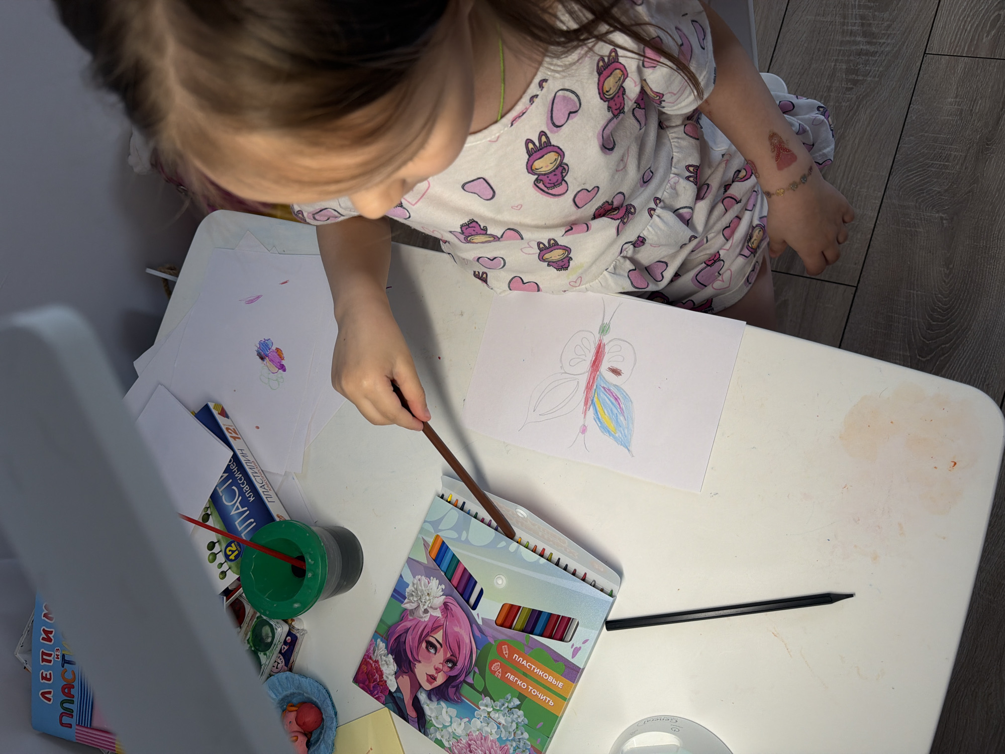A child is sitting at a white table, drawing a butterfly with colored pencils. The child has brown hair and is wearing a white dress with pink bunny designs. There is a box of colored pencils with an anime-style girl printed on it, a green pencil holder with a brush, a book titled "Пластилин" (Plasticine in Russian), and other art supplies on the table. The location is identified through text on a box in Russian: Chaikovsky, Russia.