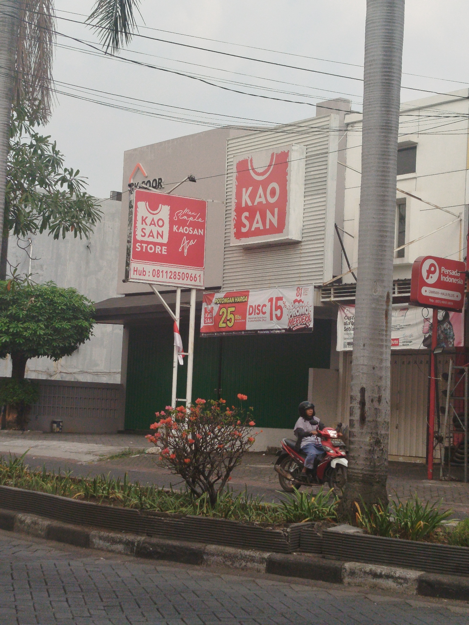 A street scene in Yosowilangun, Indonesia, shows a building with several signs. One sign, in red and white, reads "KAO SAN STORE." Another red sign, with a white "P" logo, indicates "Persada Indonesia U MRAH HAJJ PLUS." A man wearing a helmet is riding a red motorcycle on the street. In the foreground, there is a sidewalk with plants and a paved road. The sky is overcast.