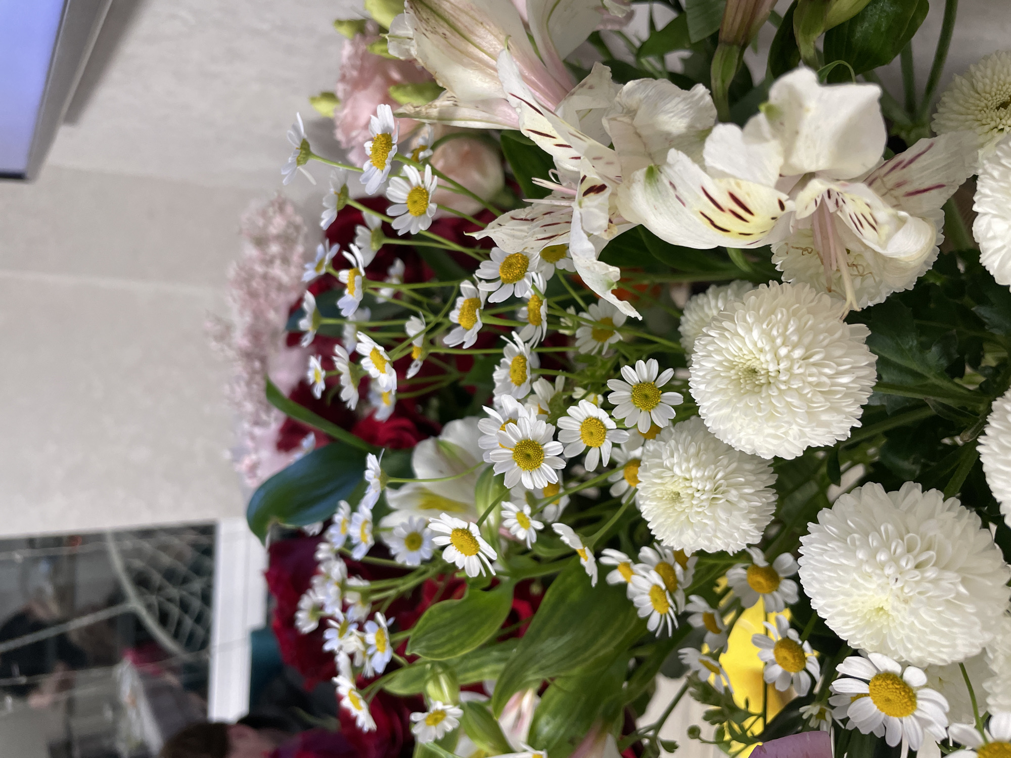 The image depicts a bouquet of flowers. The primary flowers include white daisies with yellow centers, large white pompom chrysanthemums, and white alstroemeria with pink streaks. There are also red roses and pink filler flowers. The bouquet is arranged with green leaves. The background is a blurry mix of an interior setting, possibly a room with a reflection of windows and a partial view of a television screen. The location is not discernible as Izhevsk, Russia based on the image alone.