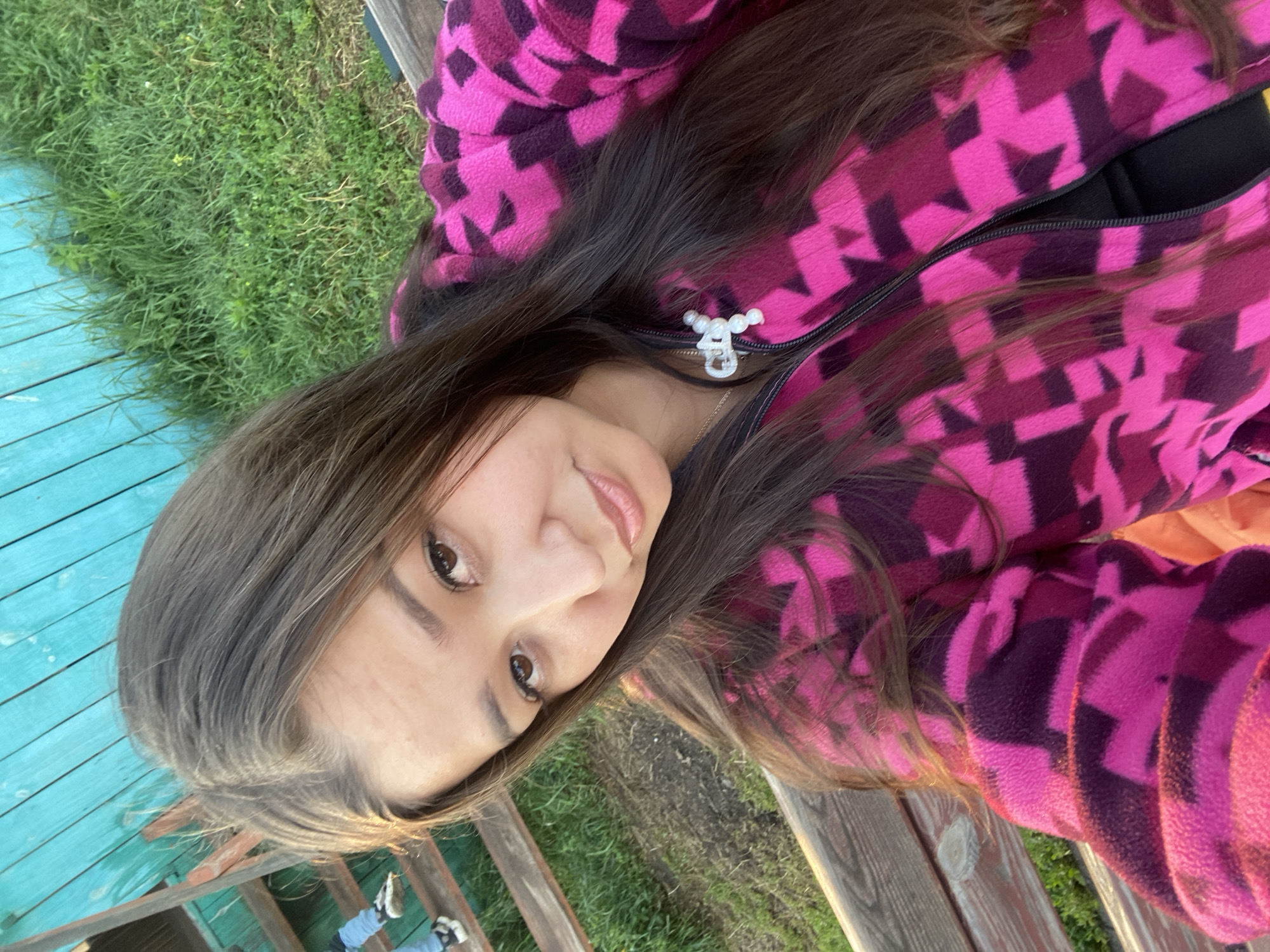 In the city of Maloarslangulovo, Russia, a young woman with long, dark brown hair is pictured in a close-up selfie. She is wearing a vibrant pink fleece jacket with a geometric purple pattern. The jacket has a zipper front, and a delicate necklace with a pearl charm is visible. Her eyes are brown, and she has fair skin with a light tan. The background features a weathered wooden fence painted turquoise, lush green grass, and a wooden bench. Sunlight casts a warm glow on the scene, suggesting it is late afternoon. In the upper left corner, a wooden staircase and a glimpse of blue jeans on a person descending are visible.
