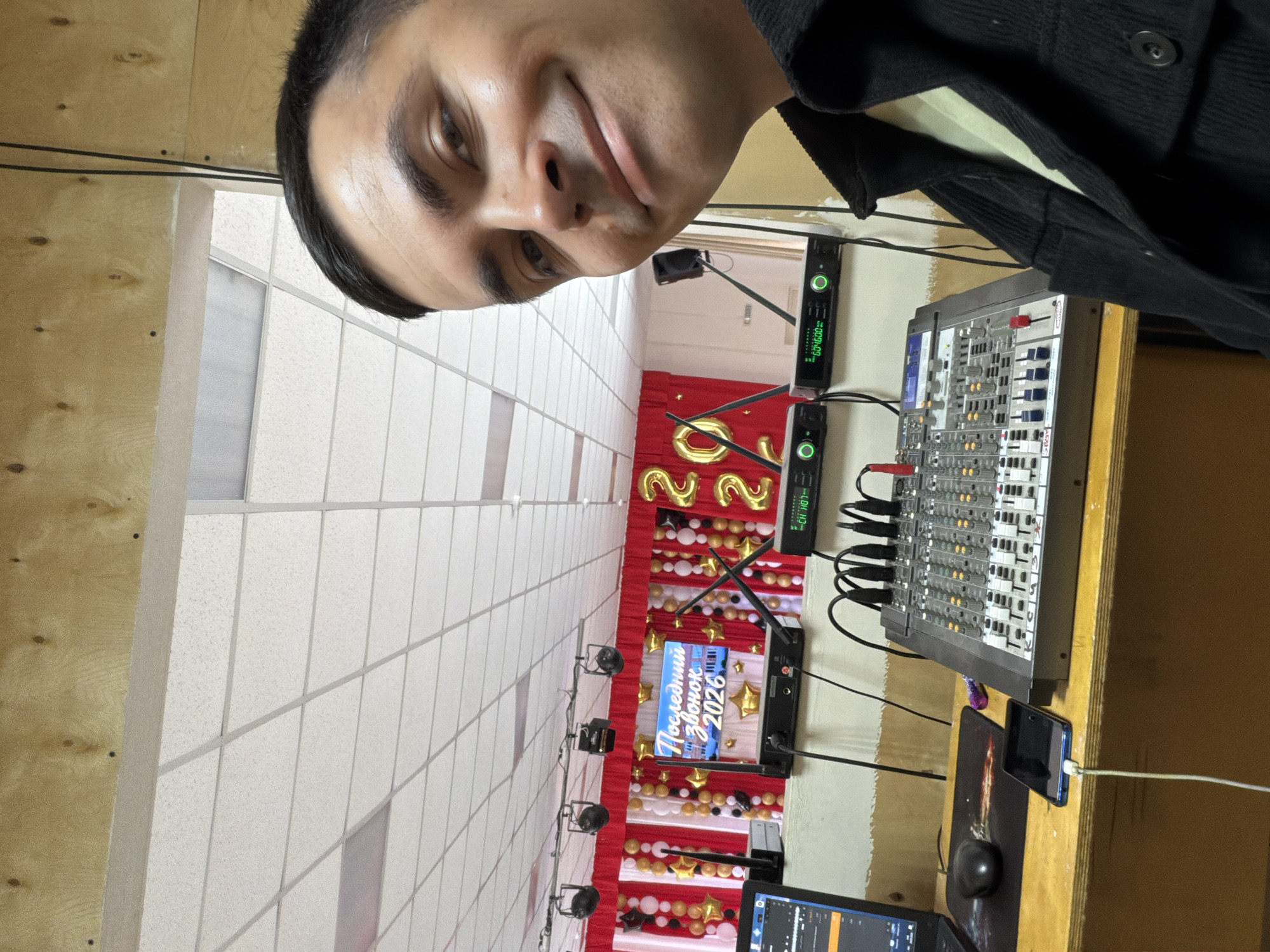 A man takes a selfie in a room. The man has dark hair and eyes, and light skin. He is wearing a dark jacket over a light-colored shirt. To his right is an audio mixing board on a wooden table. In the background is a red and white banner with gold balloons that say "2026" and a screen that reads "Последний Звонок 2026" ("Last Bell 2026" in Russian). The room appears to be in a school or auditorium. This image was likely taken in Izhevsk, Russia.