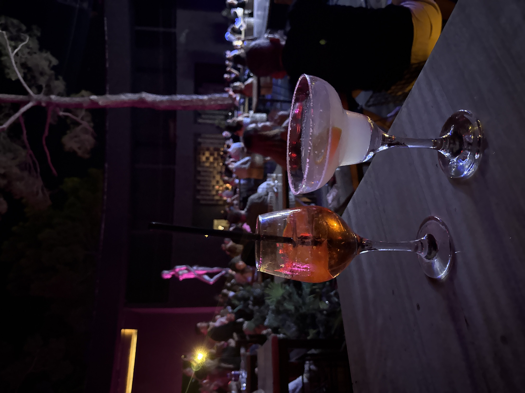 Two cocktails are on a table in the foreground. The glass on the left is a stemmed wine glass filled with an amber-colored liquid and a black straw. The glass on the right is a margarita glass with a sugar-rimmed rim, filled with a pale pink liquid and garnished with citrus slices. In the background, a crowd of people are seated at tables in an outdoor venue with purple and pink lighting. A statue of a person is visible in the background, illuminated by pink light. The scene appears to be a bar or restaurant in Bodrum, Turkey, at night.