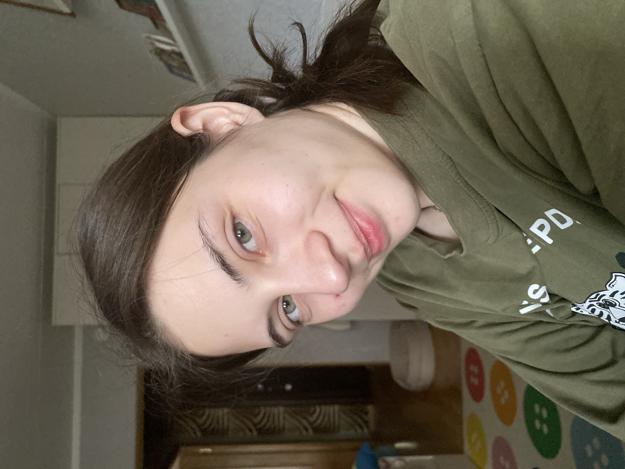A young woman with green eyes and dark brown hair, partially pulled back, is wearing a green t-shirt with white lettering and a graphic design. She is looking at the camera with a slight smile. The background shows a room with light-colored walls, a dark wood cabinet, and a floor with a colorful button-themed rug. There is insufficient information to accurately identify if the image was taken in Votkinsk, Russia, or anywhere in Russia.