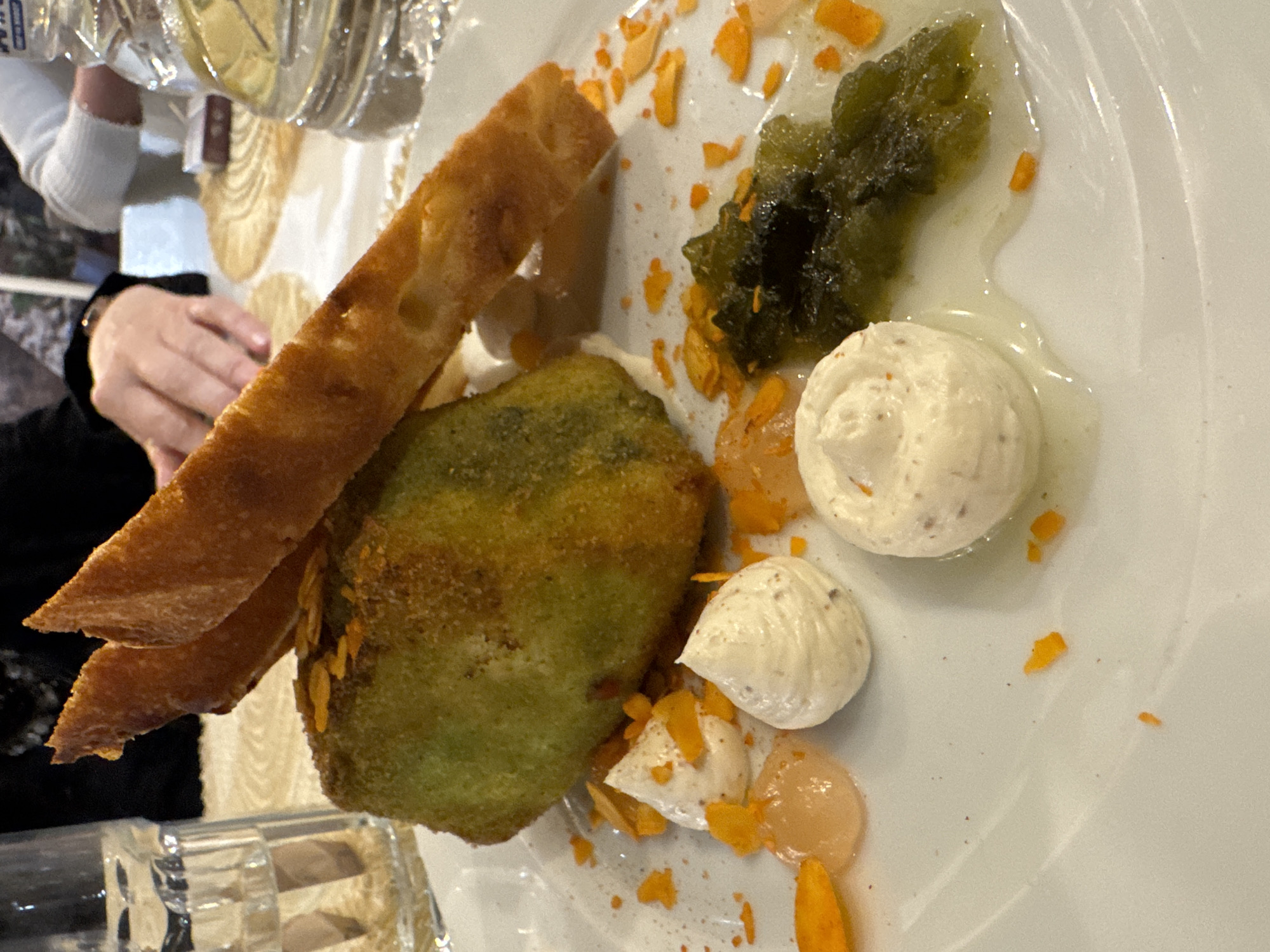 A plate of food is displayed at the restaurant "Rose" in Pomorie, Bulgaria. The dish features a green, oblong item, possibly a piece of food, coated in crumbs and topped with slivers of toasted bread. Surrounding it are dollops of white cream, a translucent orange gel, and a dark green, gel-like substance. Orange flakes are scattered across the plate. In the background, a glass of water and a bottle of water are visible. Two people are present, with one's hands visible in the upper center of the frame.