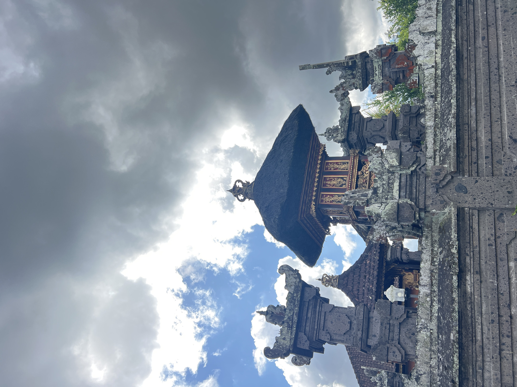The image depicts a traditional Balinese temple complex, likely located in Kintamani, Indonesia, as evidenced by the architectural style. The foreground features a textured stone wall, with intricate carvings and decorative elements suggestive of ancient craftsmanship. Above the wall, ornate stone pillars and archways rise, adorned with moss and lichen, indicating age and exposure to the elements. Dominating the midground is a tiered pagoda-style structure with a thatched roof and intricately decorated golden panels. The sky above is dramatic, with a mix of dark, heavy storm clouds and patches of bright blue sky, suggesting a dynamic weather system. The overall scene conveys a sense of cultural heritage and natural grandeur.