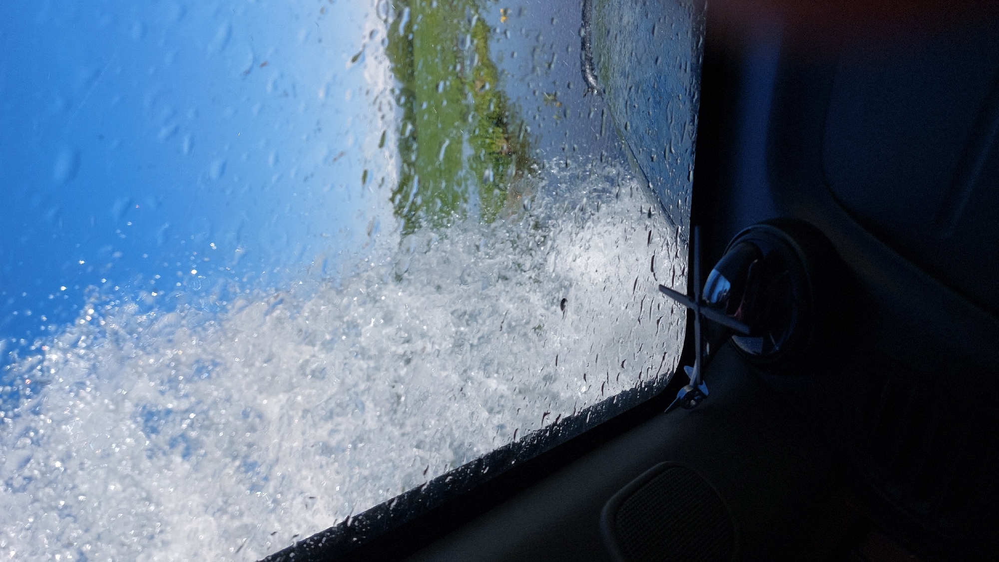 The image depicts a view from inside a vehicle, looking out through the windshield. Large splashes of water obscure a significant portion of the view. Beyond the water, a bright blue sky is visible, along with a green, watery landscape that appears to be a flooded field or a body of water with vegetation. The dashboard of the vehicle is partially visible in the foreground, with a small airplane-shaped ornament on a circular base mounted on it. The scene suggests a vehicle driving through or encountering a substantial amount of water. There is no indication of the city of Koryaki, Russia in the image itself.