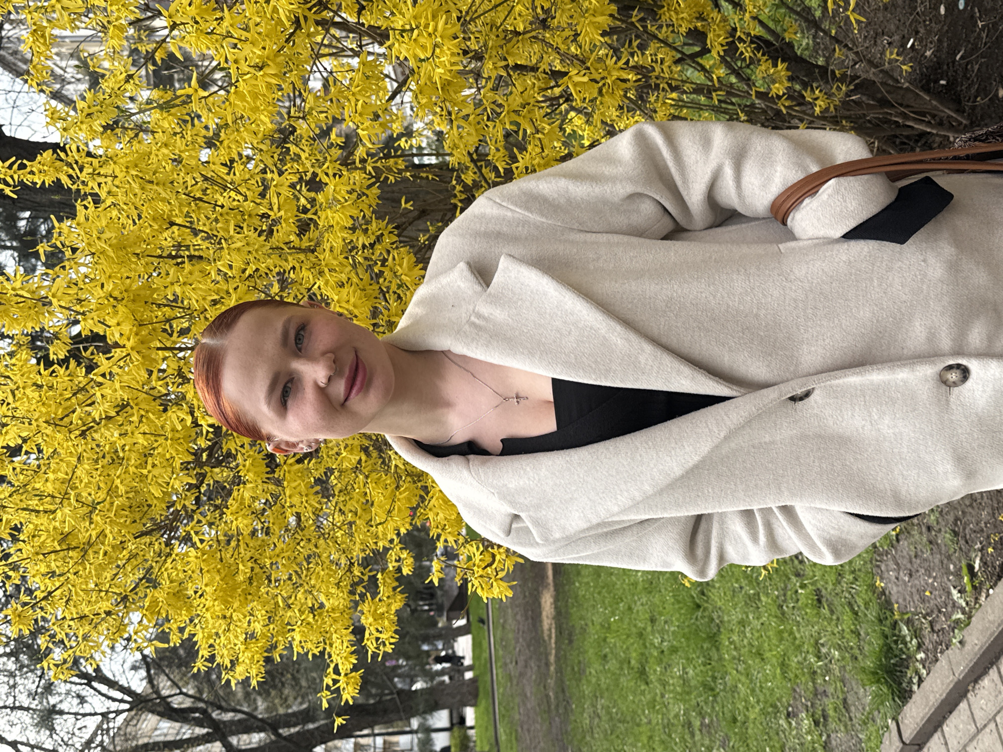 A young woman with red hair is standing outdoors next to a blooming forsythia bush filled with bright yellow flowers. She is wearing a beige coat over a black top and a necklace. The environment includes green grass, trees, and a sidewalk, suggesting a park or garden setting. The location of Ovoshchnoy, Russia, cannot be verified from the image.
