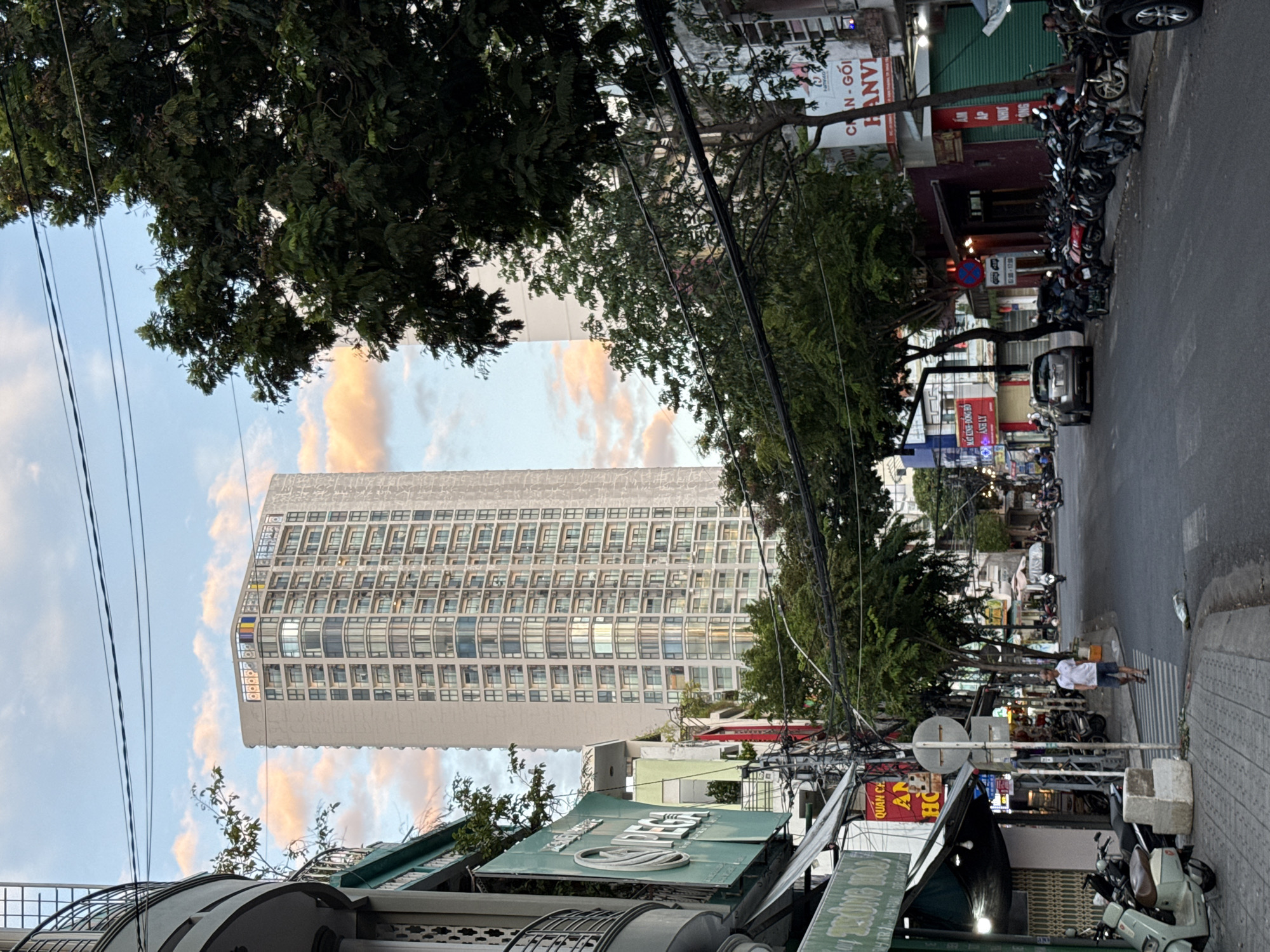 The image depicts a street scene in Nha Trang, Vietnam. A tall, modern apartment building with numerous balconies dominates the background. In the foreground, a street is lined with trees and buildings, many of which appear to be shops or businesses, indicated by signs with Vietnamese text. Various motorbikes are parked along the street and sidewalk. A lone man in a white shirt and shorts walks across a pedestrian crossing. The sky is partly cloudy with hues of pink and orange, suggesting it might be during sunrise or sunset. The overall atmosphere is that of a busy urban street in Southeast Asia.