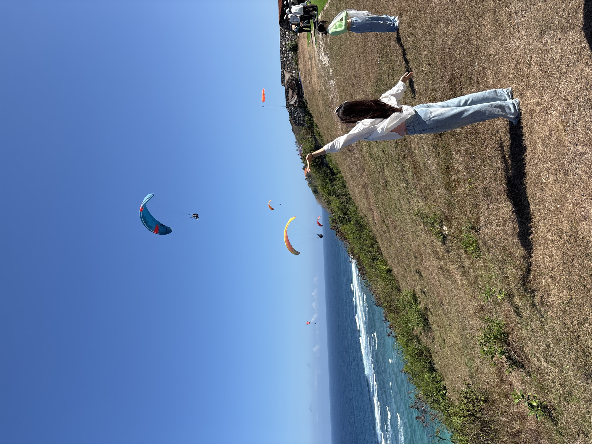 The image depicts a paragliding site near the coast in Kuta Selatan, Indonesia. In the foreground, a woman with long dark hair, wearing a white open shirt over a pink top and light blue jeans, stands with her arms outstretched, facing away from the camera and towards the ocean. To her right and further back, another person in a light green top and blue jeans stands. Several paragliders are visible in the clear blue sky over the turquoise ocean. The landscape consists of a grassy hill with patches of dry vegetation leading down to the sea, where waves are breaking. In the background, a stone wall structure is visible, along with a group of people and a flag. The scene suggests an outdoor recreational activity in a scenic coastal location.