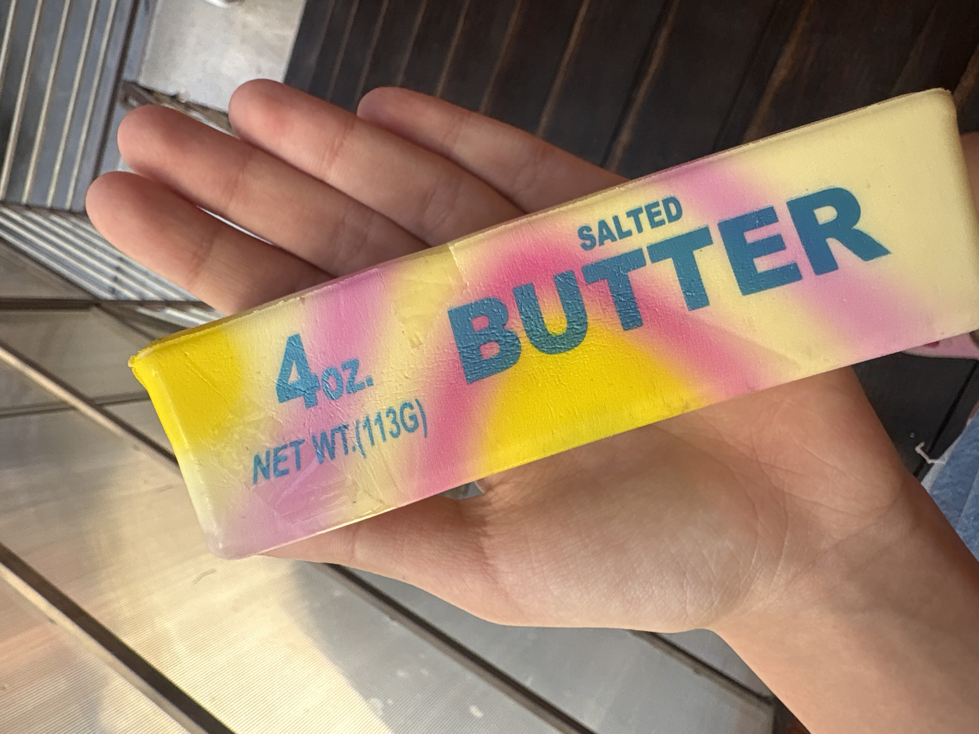 A person holds a stick of butter with a pastel tie-dye wrapper in their hand. The butter stick is yellow, pink, and purple. Text on the wrapper reads "4 OZ. NET WT. (113G)" and "SALTED BUTTER". The background is out of focus but appears to be an outdoor setting with a textured metal surface and dark wooden planks. The image was taken in Georgiyevsk, Russia.