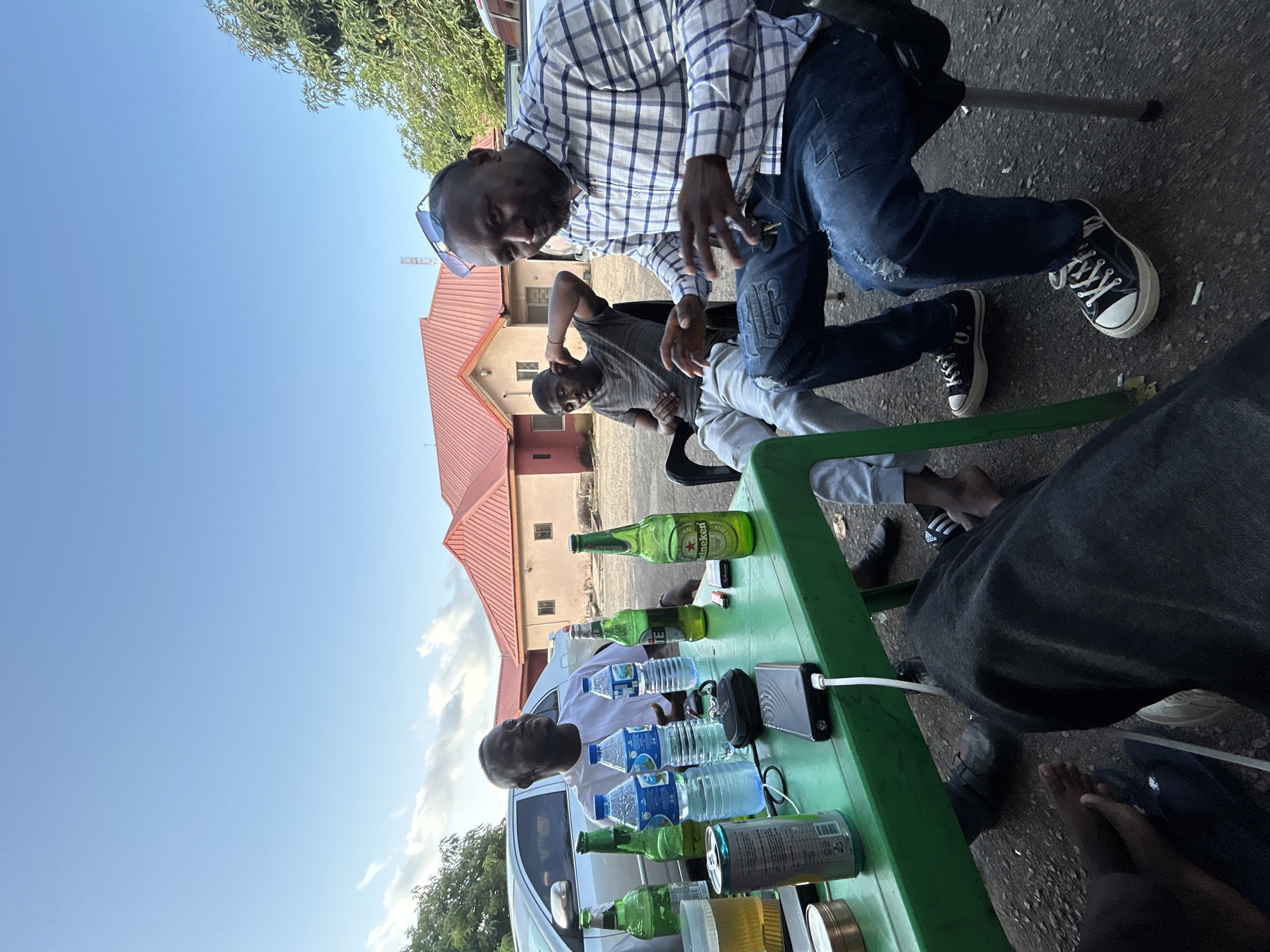 Four dark-skinned men are gathered outdoors around a green plastic table in Jos, Nigeria, during daylight hours under a clear blue sky with sparse clouds.
**People:**
* **Left man:** Wearing a white short-sleeved top, seated at the left side of the table, looking towards the right with a slight smile.
* **Center-back man:** Wearing a gray t-shirt and light gray sweatpants, seated on a black chair behind the table. His left hand is raised, touching his ear. His feet are bare.
* **Right man (foreground):** Wearing a blue and white plaid long-sleeved shirt and distressed blue jeans, with blue-tinted sunglasses resting on his head. He is seated on a black office-style chair, looking towards the camera with his hands raised and gesturing. Keys are clipped to his jeans. He wears black canvas sneakers with white soles and laces.
* **Bottom-left man (partially visible):** Only his legs and feet are visible under the table. He wears dark pants, and one foot is bare, while the other wears a dark sandal with a white Adidas logo.
**Objects on the Table (Green Plastic):**
* Several green glass bottles, including one Heineken and one "33" Export Lager Beer.
* Three clear plastic bottles of "SIA Water."
* A silver aluminum beverage can.
* A clear glass mug containing a yellow liquid (presumably beer) with foam.
* A black power bank with a white USB charging cable connected.
* A black smartphone or similar device next to the power bank.
* A small black lighter.
* A pack of cigarettes.
* A small circular metallic tin.
**Environment:**
The scene is set on an uneven concrete or dirt surface. In the background, a single-story building with a prominent reddish-orange corrugated metal roof and light-colored walls is visible. A silver sedan is partially parked behind the left man. Green trees and foliage are present on both sides of the frame. A telecommunications tower is faintly visible in the distant background.