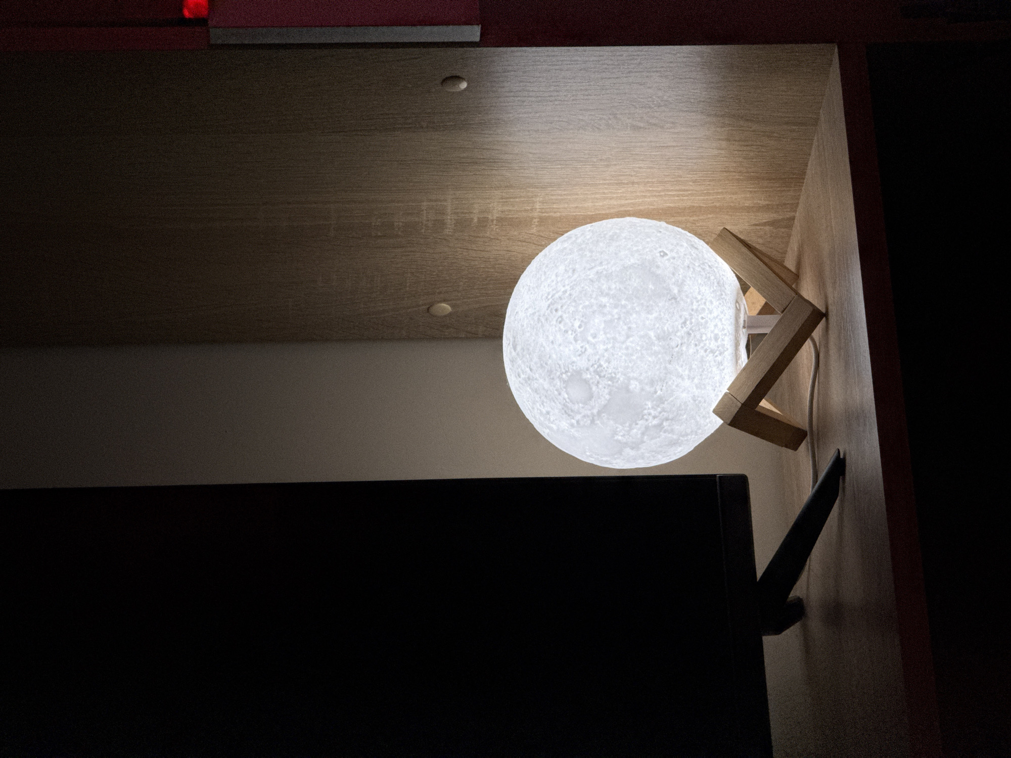 The image displays an indoor scene in low light conditions, located in Unknown, Unknown. A spherical, illuminated lamp with a textured surface resembling the moon rests on a light-colored wooden tripod stand, occupying the central-right portion of the frame. A white power cord is visible near the base of the lamp. To the left, a section of a dark, unlit electronic display (likely a monitor or television) with a black stand is present. All objects are situated on a flat, light brown wooden surface, consistent with a desk or shelf. Behind the moon lamp and to its right, a vertical light brown wooden panel, part of a furniture unit, features two circular, lighter-toned plugs or caps. A dark red vertical surface is partially visible along the far right edge, exhibiting a small, bright red illuminated indicator light in the upper right corner. No people are present.