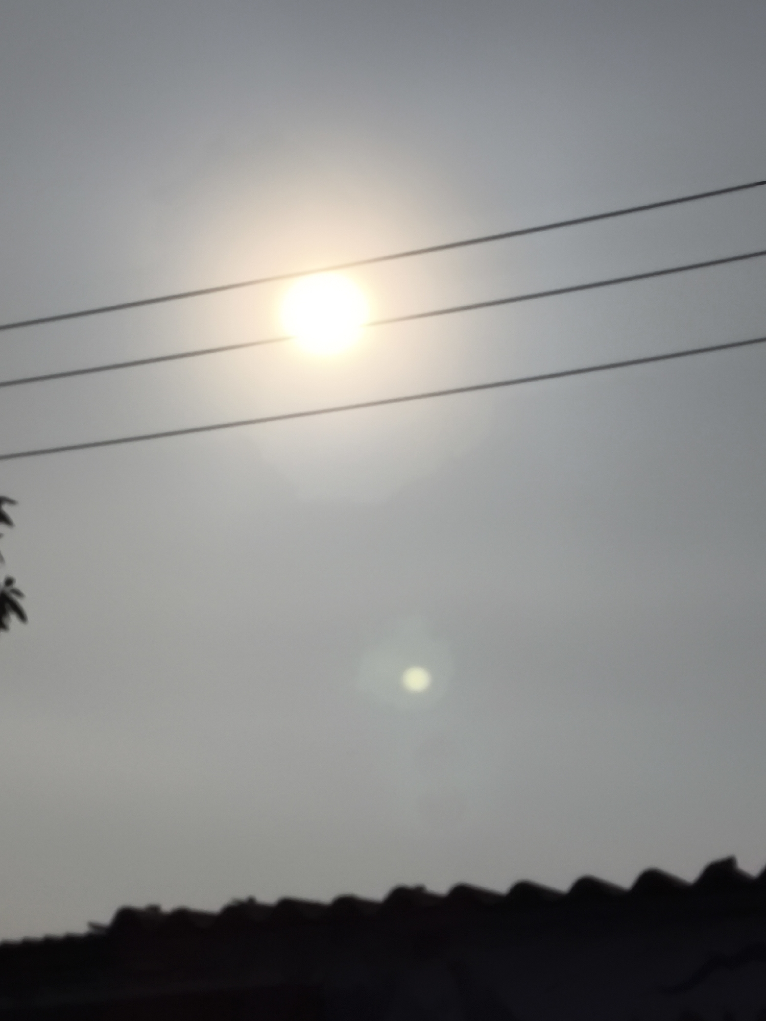 The image depicts a hazy sky with the sun partially obscured by atmospheric conditions, creating a soft glow. Three parallel black wires, likely power lines, stretch horizontally across the upper portion of the frame. In the lower section, a silhouetted structure with a jagged, dark outline suggests the roof of a building, possibly a house or a series of dwellings. A faint lens flare or atmospheric light artifact is visible below the sun. The overall scene conveys a muted, overcast atmosphere. The image does not provide specific clues to definitively pinpoint its location within Cilegon, Indonesia, other than it appears to be an outdoor scene with visible electrical infrastructure and residential rooftops.