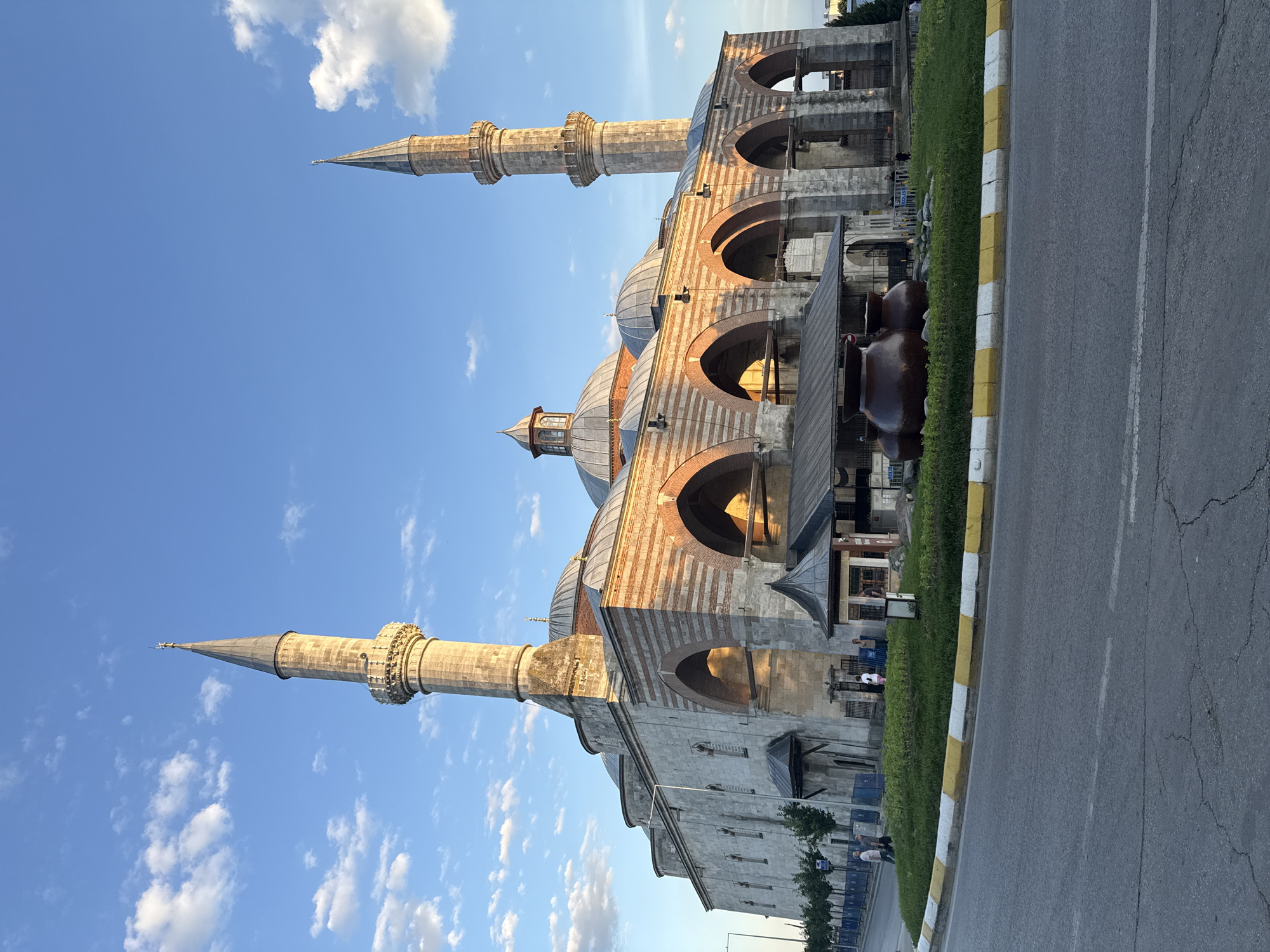 A wide-angle, eye-level shot captures the Selimiye Mosque in Edirne, Turkey, under a bright blue sky with scattered white clouds. The mosque, a UNESCO World Heritage site, is a prominent example of Ottoman architecture. Its grand stone structure features multiple domes, the largest being at the center, and two slender, tall minarets flanking the building. The lower section of the mosque exhibits striking red and white striped arches. In front of the mosque, a circular green median is adorned with three large, dark brown ceramic pots. A curved asphalt road with yellow and white markings is visible in the foreground. Several individuals are casually walking near the entrance of the mosque, and a few are standing on the sidewalk on the left side of the image. The scene is illuminated by the warm glow of the late afternoon sun.