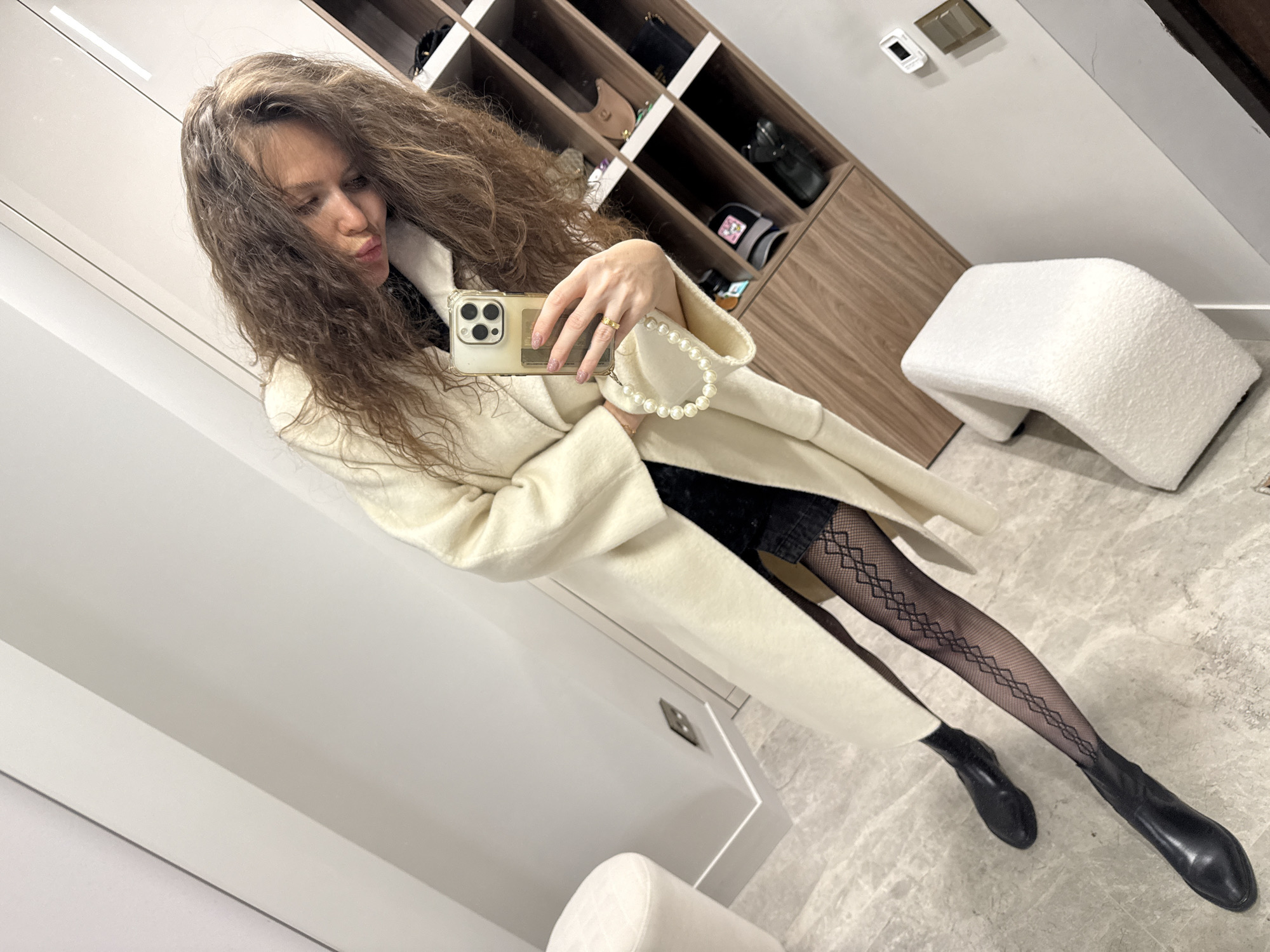 A woman with long, wavy brown hair is posing for a selfie in what appears to be a walk-in closet. She is wearing a white coat, black textured stockings, a short black skirt, and black boots. She is holding a white iPhone with a pearl wrist strap in her left hand, while making a kissing face toward the camera.
Behind her is a closet system with open shelves containing various items, including handbags and hats. To her right, a curved white ottoman sits on a gray marble floor. The walls are primarily white, and there is a light switch visible above the closet system.
The location is not discernible from the image; no information about the city of Khimki, Russia, is discernible.