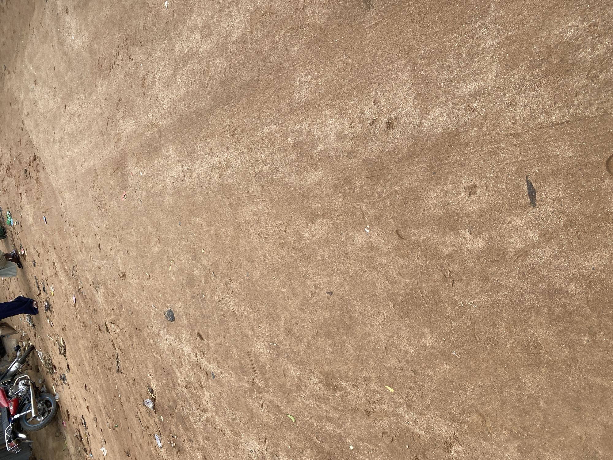 The image depicts a dusty, brown, uneven surface, likely a dirt road or clearing, with some debris scattered across it. In the lower left portion of the image, a red motorcycle is visible, parked on the ground. To the left of the motorcycle, there are glimpses of people, with one person wearing a blue garment and another in darker clothing. The scene appears to be outdoors. The lighting suggests it is daytime, with the sun casting some shadows and highlighting the texture of the ground. There is no discernible text in the image. The overall impression is of an unpaved, possibly rural or less developed area, given the presence of the motorcycle and the natural, unkempt ground. The scattered litter indicates a lack of regular maintenance.