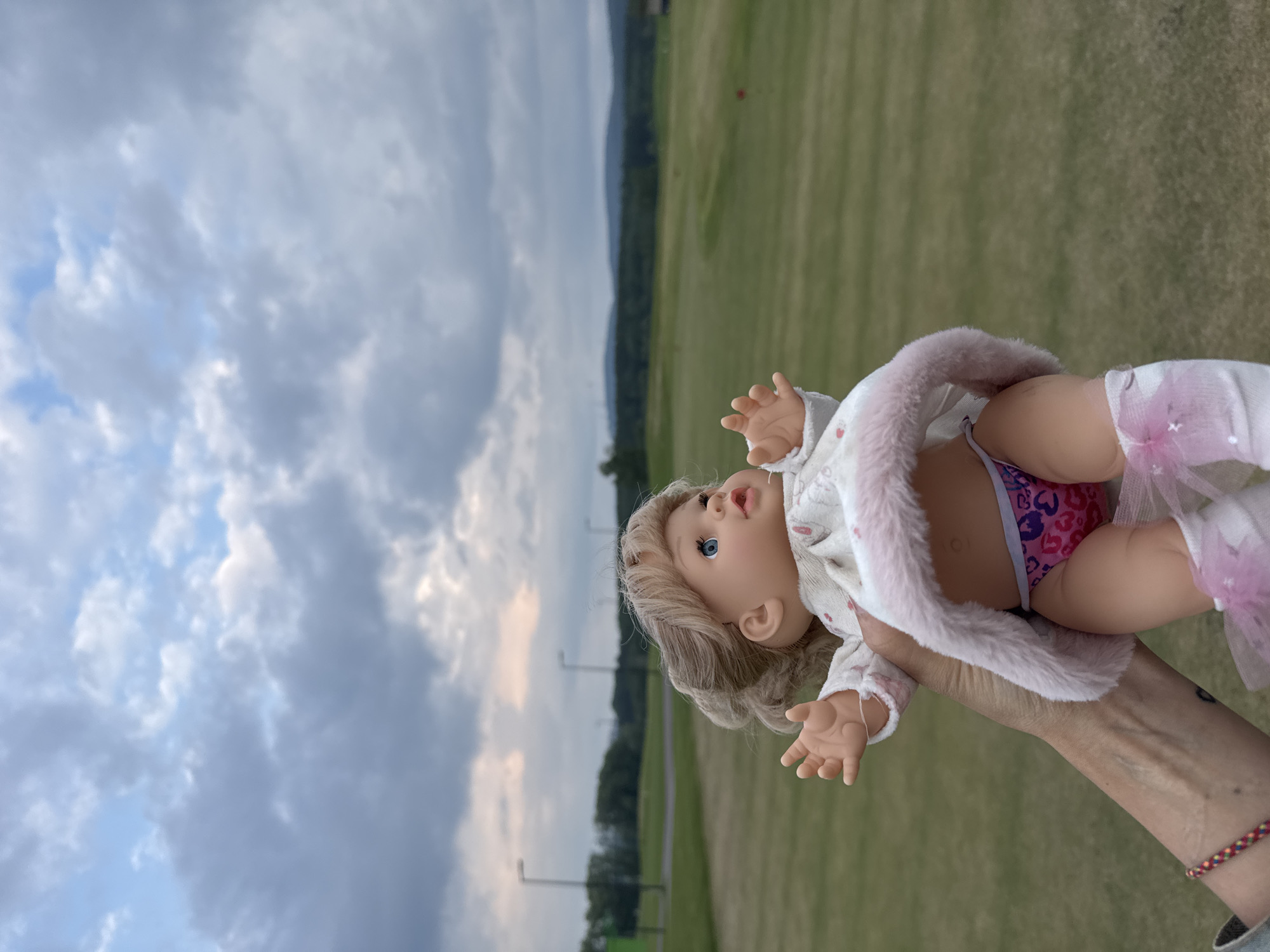 An adult's hand holds a baby doll in front of a green grassy field with distant trees and a cloudy sky. The doll has blonde hair, blue eyes, and is wearing a white dress with pink trim, pink underwear with hearts, and pink socks. The hand has a red, yellow, and green bracelet on the wrist. The background appears to be a golf course. This image was taken in Krasnoyarsk, Russia.