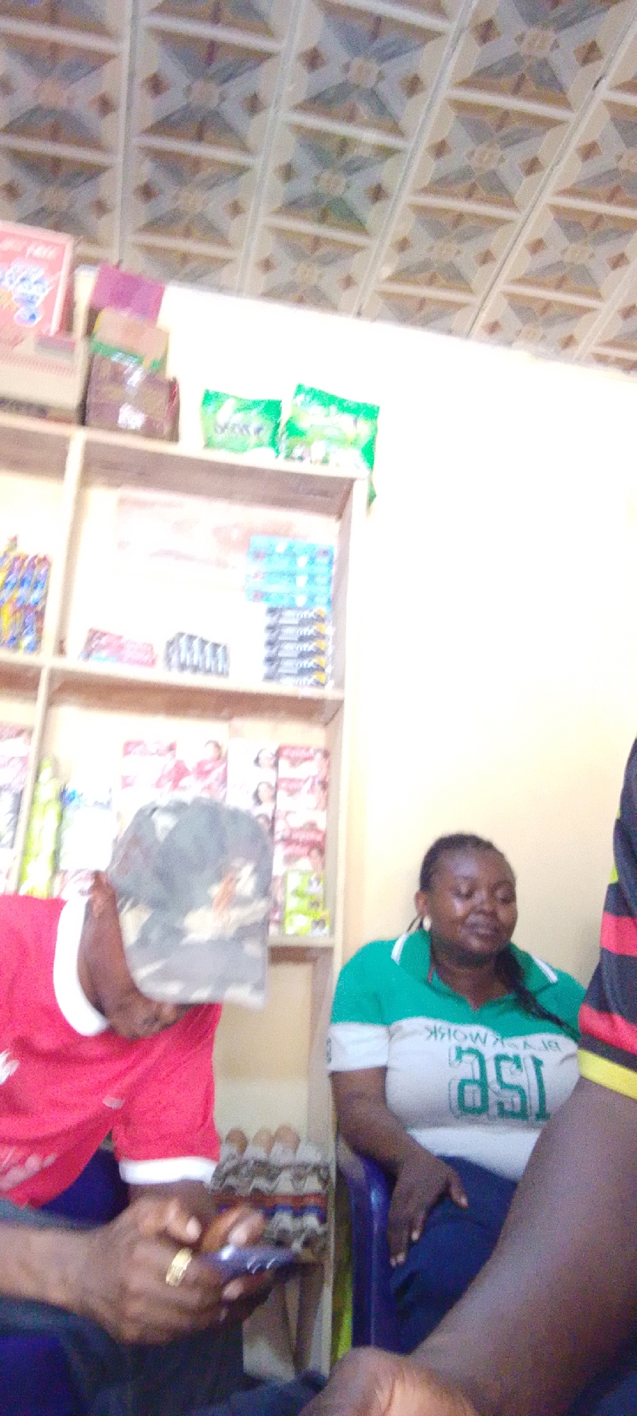 Three people are in what appears to be a small store in Adankolo, Nigeria. A man in a red shirt and camouflage hat is looking at a phone while holding a carton of eggs. A woman in a green and white shirt is seated. Part of another person wearing a red, black, and yellow striped shirt is visible on the right. Shelves stocked with various products are visible in the background, and the ceiling has a tiled pattern.