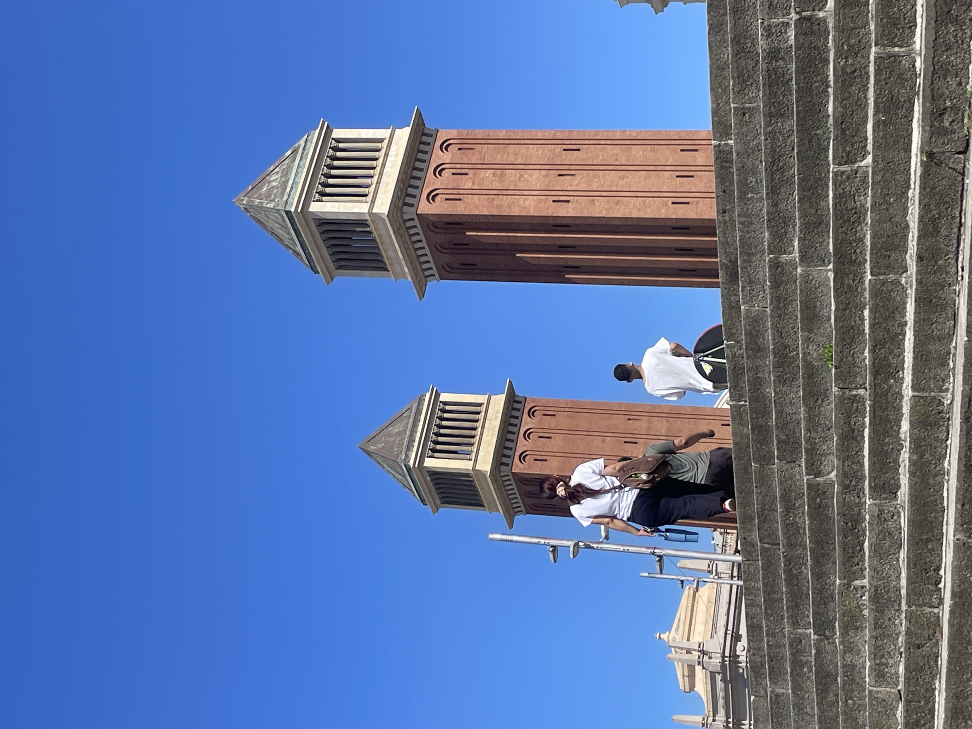 The image captures an outdoor scene in Barcelona, Spain, showcasing two prominent architectural towers set against a clear, brilliant blue sky. These are the iconic Venetian Towers, distinguished by their reddish-brown brick bodies and elaborate, light-colored stone upper sections featuring columns and pyramidal copper-patinated roofs.
On the right side of the frame, a wide, grand stone staircase ascends, constructed from large, textured gray blocks. Several individuals are present on these steps. In the foreground, a person is seen wearing a white t-shirt, dark pants, and carrying a brown messenger bag, with a face mask visible under their chin. They are standing near a metal railing or scaffolding. Further up the steps, another person in a white t-shirt and dark clothing is partially visible.
The vibrant blue sky indicates a sunny day, likely midday or early afternoon, with strong natural light illuminating the scene. The weather appears clear and pleasant. A small section of another ornate, light-colored building structure can be glimpsed below the main staircase on the far left, suggesting the broader architectural environment of the location. There is no visible text in the image.