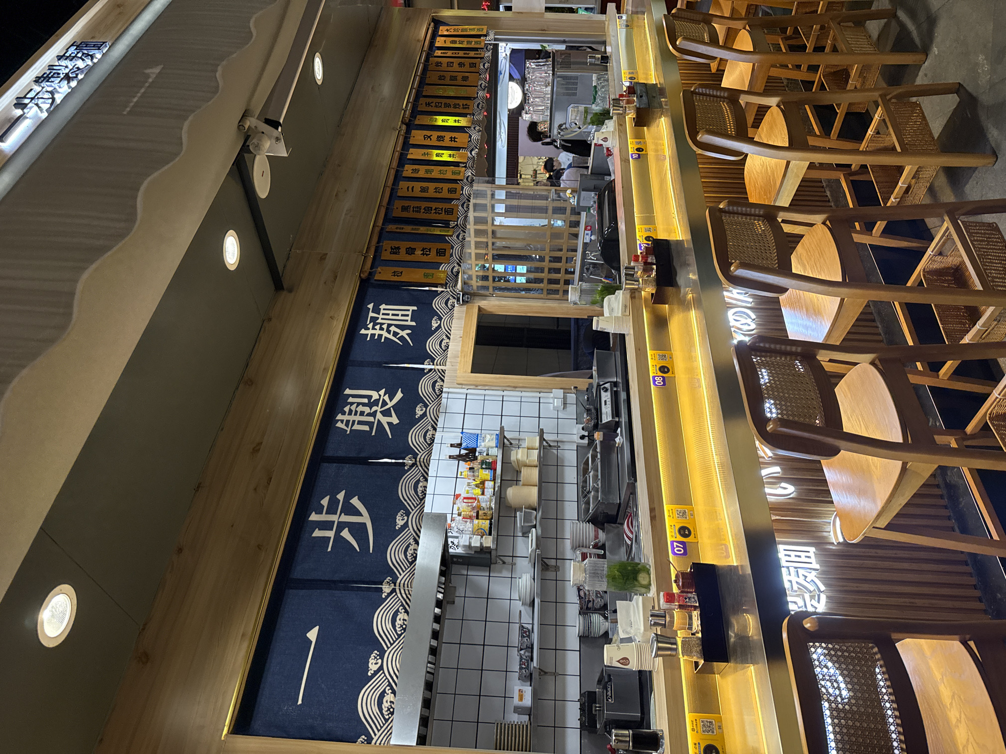 This is an interior shot of a restaurant, likely located in Haizhu District, China, based on the Mandarin signage.
The restaurant features a counter with high wooden chairs. There are menus with names of dishes in Chinese and Japanese, arranged vertically on the wall. A blue curtain with white Chinese characters and wave designs partially obscures the view into the kitchen. The kitchen area is visible behind the counter, revealing shelves stocked with various ingredients and supplies. There are QR codes on the front of the counter.
The overall impression is a modern restaurant with an open kitchen design.