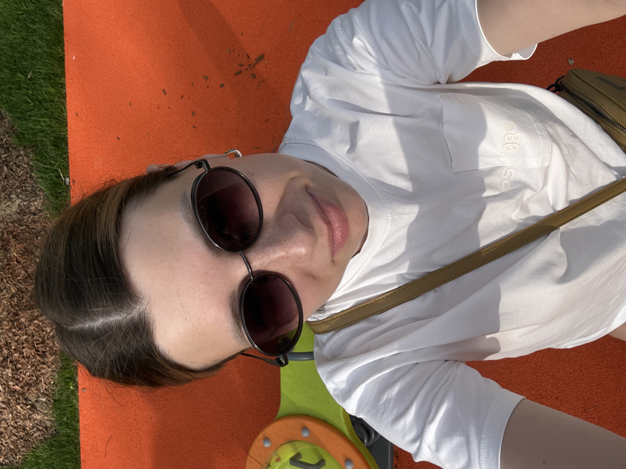 A person wearing sunglasses and a white t-shirt is lying on a red surface. A brown bag with a strap is positioned next to them. Adjacent to the red surface is an area of green grass and a patch of brown ground. In the foreground, parts of a playground structure with green and orange elements are visible. The location cannot be determined from the image.