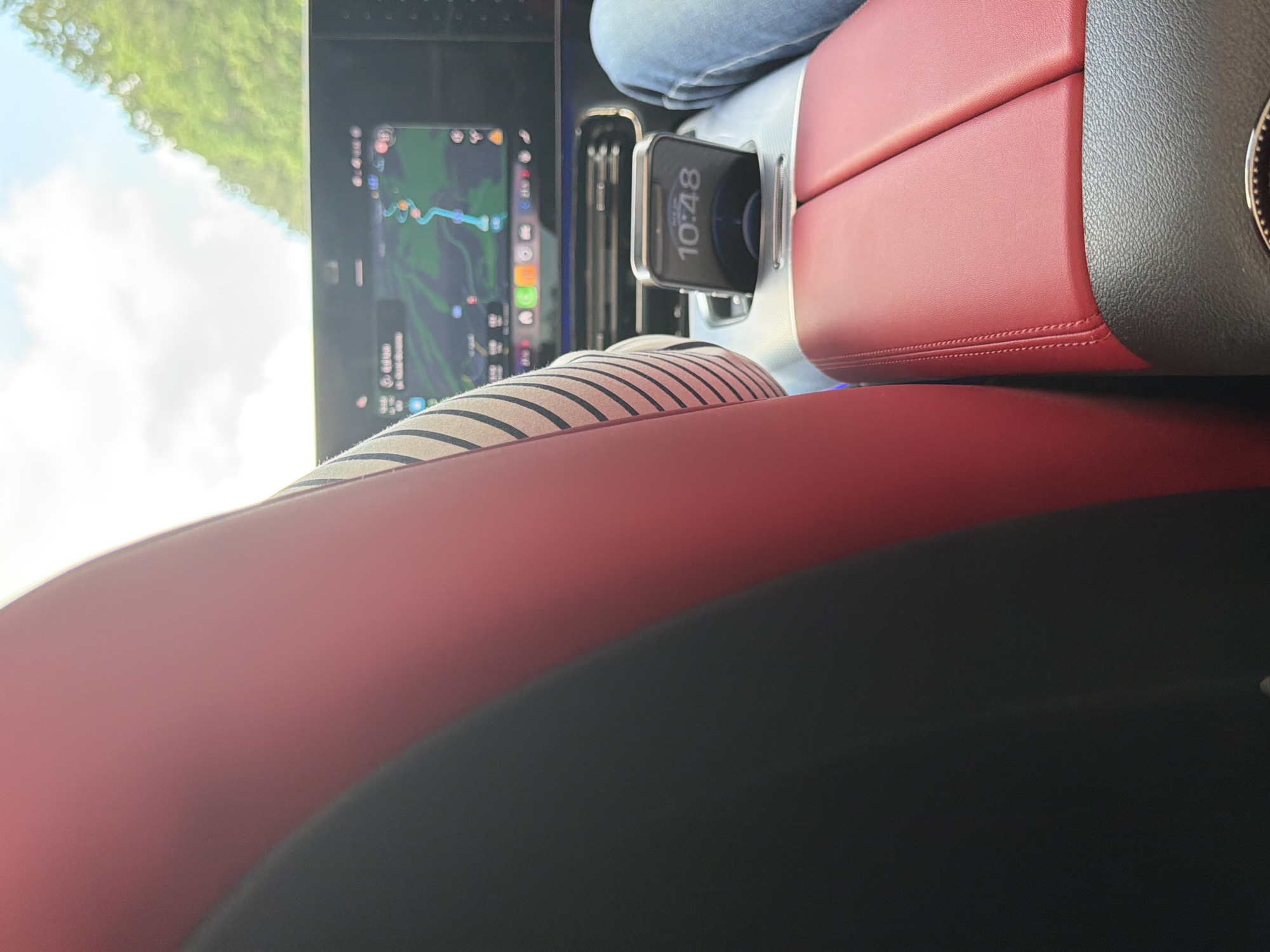 The image provides a view from within a modern car, likely from a passenger's perspective looking towards the front and right. The foreground features prominent red leather upholstery, potentially a car seat or armrest, with a distinct stitch line. Adjacent to this, a black textured surface is visible.
In the mid-ground, a person's lap is partially visible, clad in blue jeans and a horizontally striped fabric, possibly part of their attire or a blanket. Situated within a light gray or silver center console, a smartphone displays the time "10:48" on its screen, appearing to be in a charging or holding dock. To the left of the console, a large infotainment screen is active, showing a navigation map with a highlighted route. Several app icons, including those for music, phone, and messages, are visible along the bottom edge of this screen. Small text on the navigation screen appears to show an estimated time around "6:30."
Beyond the vehicle's interior, through a window on the left, a bright, partly cloudy sky and blurred green foliage are visible, indicating it is daytime. The overall impression is of a journey underway in a well-appointed vehicle, potentially in Borovo, Bulgaria, given the provided location context.