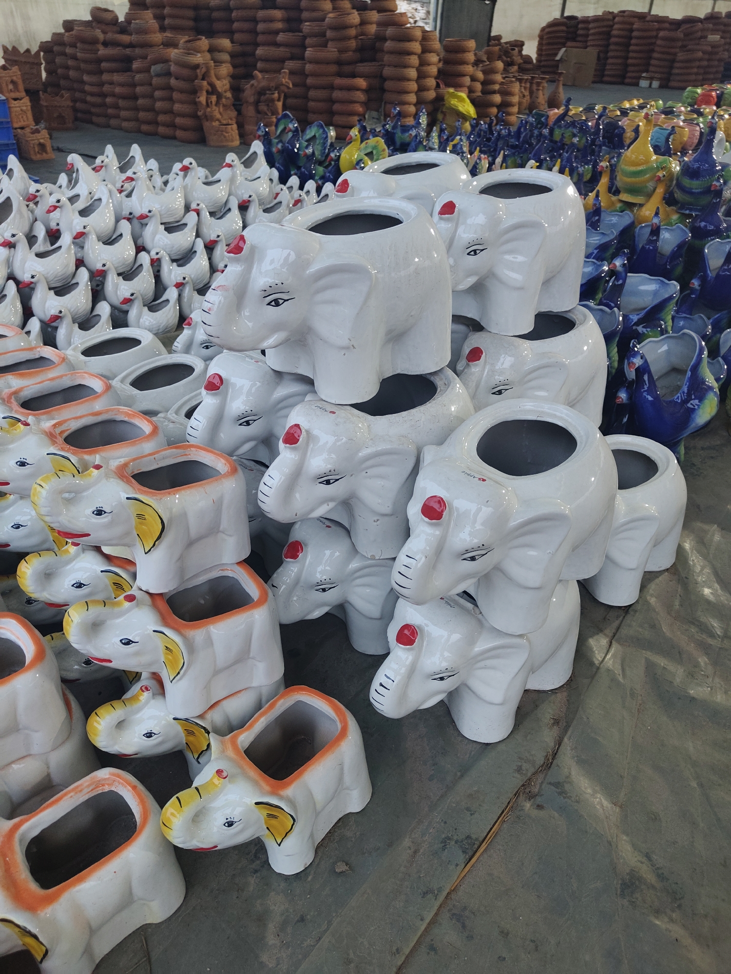 The image depicts a large assortment of ceramic pots shaped like elephants and ducks, likely for sale, in what appears to be a warehouse or large shop. The pots vary in color and design. Behind these are stacks of earthen pots and ceramic birds. The location is near ಚಂದ್ರಪ್ಪ ವೃತ್ತ, India. No people are visible in the frame. The lighting suggests an indoor setting.
