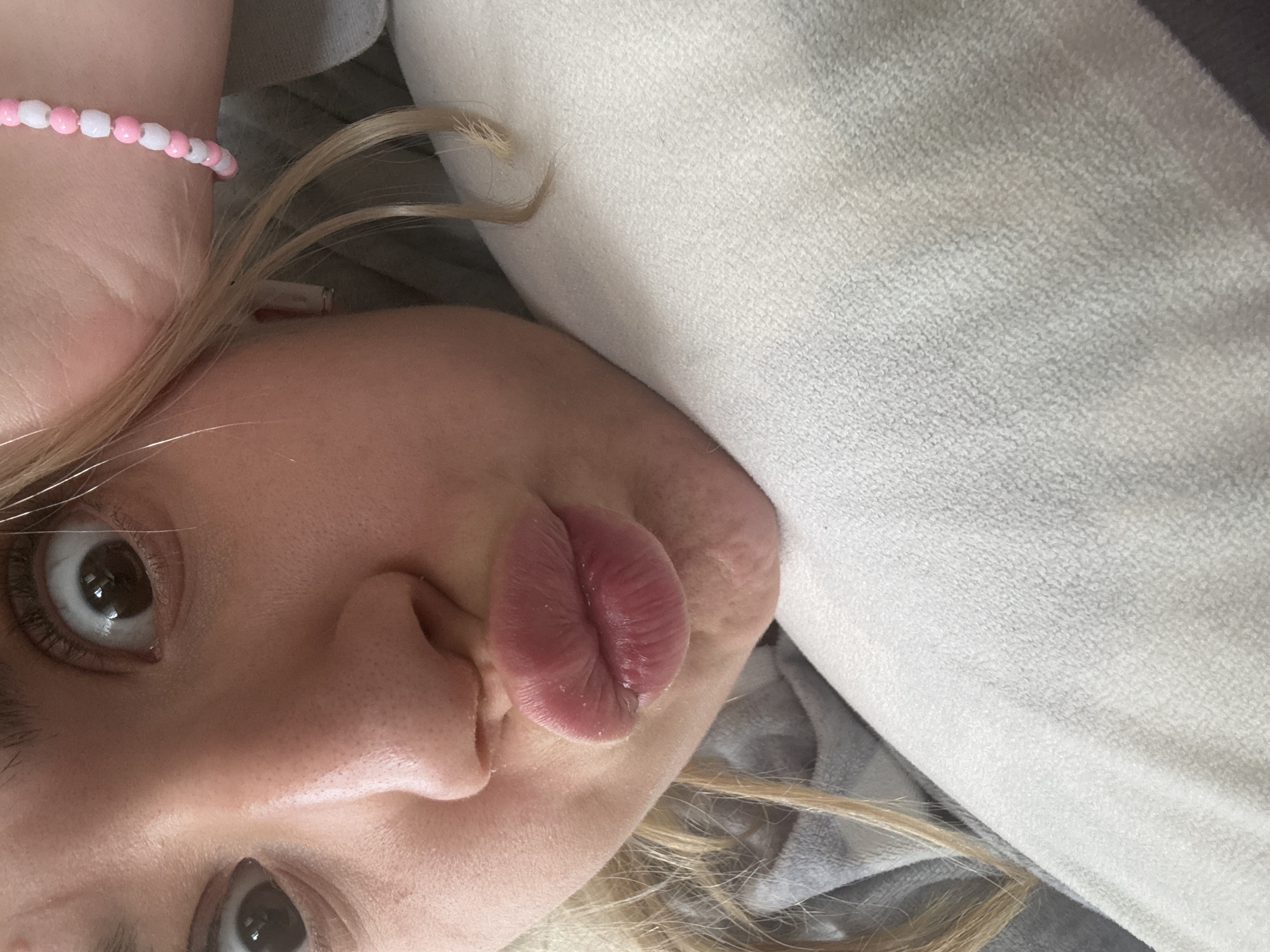 The image presents a close-up, rotated portrait of a person, likely female, captured indoors in Sofia, Bulgaria. The subject, with fair skin, blonde hair, and dark eyes, gazes directly at the viewer with a wide-eyed expression. Their lips are intentionally puckered into a "fish face" or pout, suggesting a playful or silly mood. A pink and white beaded bracelet is visible on their wrist, near the top of the frame when oriented correctly. The background features soft, textured fabrics: a prominent light-colored, possibly white or off-white, material, and a darker grey fabric, suggesting a comfortable setting like a bed or sofa. The lighting is soft and even, typical of indoor daylight. There is no visible text.