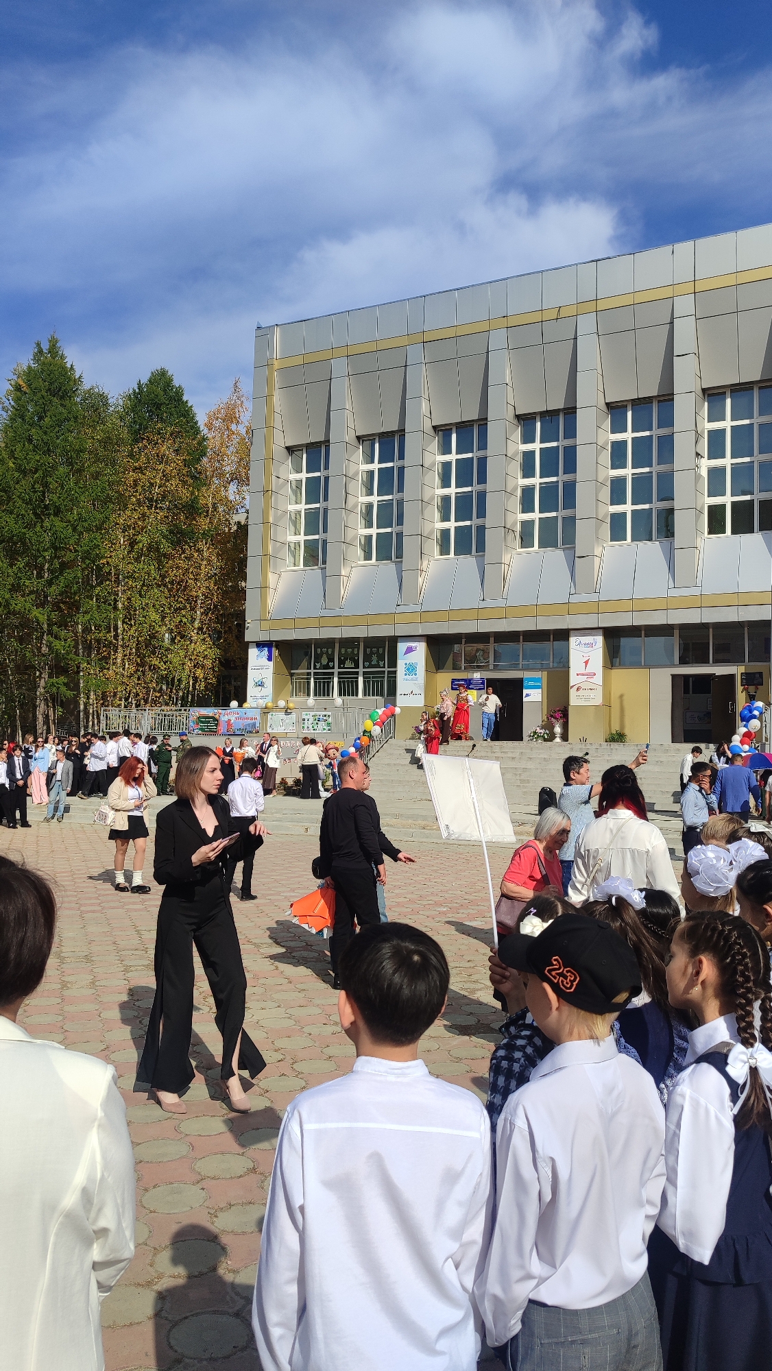 The image depicts an outdoor gathering in front of a modern building in Mirny, Russia. The building has a gray facade with a prominent yellow stripe and numerous grid-patterned windows. In front of the building, a group of people are assembled on a paved courtyard with circular stone patterns. Several individuals, including children dressed in school uniforms with white shirts and dark skirts or trousers, are visible. Some are looking towards the building's entrance, where a set of stairs leads up. Balloons in various colors, including red, yellow, green, and blue, are present, suggesting a festive occasion. Some adults are standing and conversing, while others appear to be observing the activity. One woman in the foreground is wearing a black suit with wide-leg pants and is holding a phone. Another individual in a black shirt is holding a white rectangular object on a pole. In the background, trees with green and yellow foliage can be seen, indicating it is likely autumn. The sky is blue with scattered clouds.