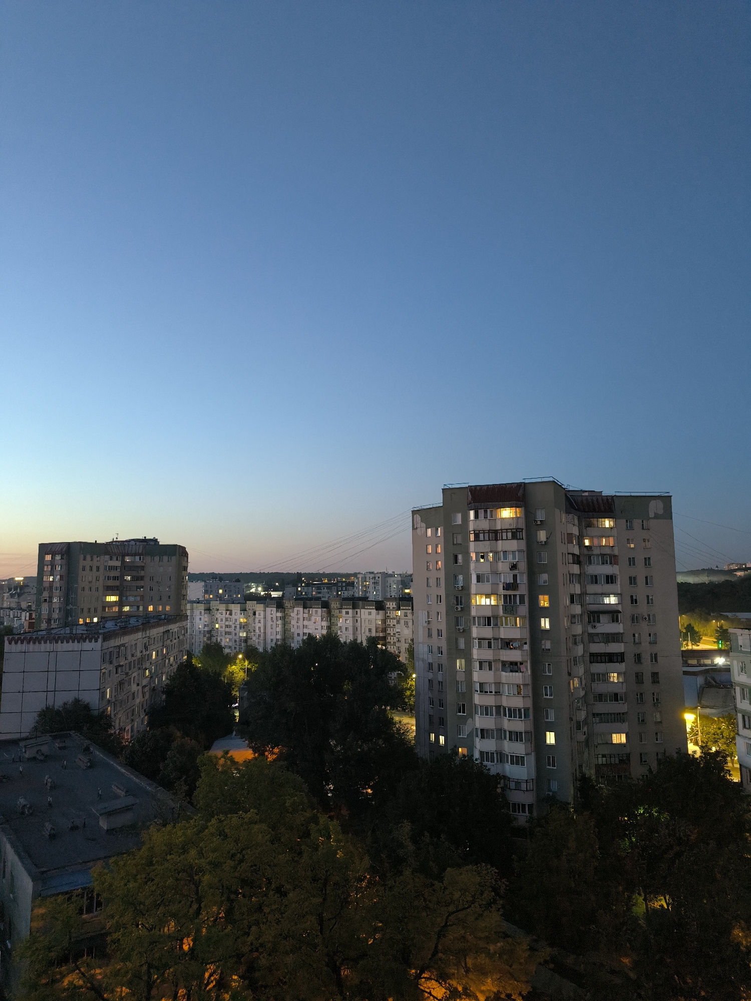 The image captures a twilight view of a residential area in Chișinău, Moldova. In the foreground, the roofs of several apartment buildings are visible, along with dense foliage from mature trees. The middle ground is dominated by several multi-story apartment blocks, characteristic of Soviet-era architecture, with lights visible in many of the windows, indicating occupancy. Some of these buildings are clad in light-colored panels, while others appear to be made of concrete. Power lines are strung between some of the structures. In the background, more apartment buildings and the silhouette of distant hills are discernible. The sky is a deep, clear blue, transitioning to a lighter hue near the horizon, suggesting the transition from day to night. There are no people visible in the image. The scene depicts a typical urban landscape during dusk.