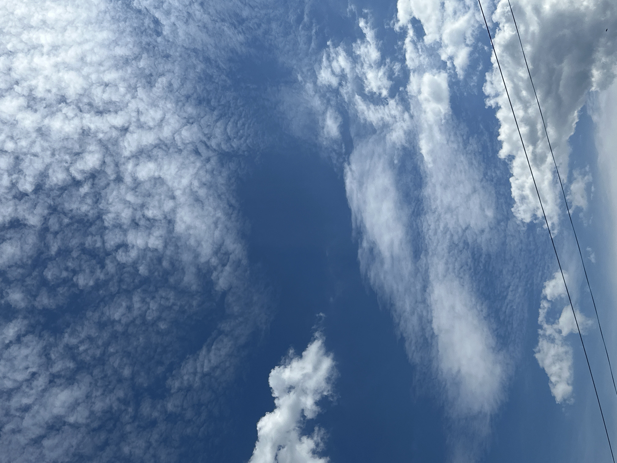 The image displays a bright, blue sky filled with various cloud formations, captured from a low vantage point looking upwards. On the left side, a significant portion of the sky is covered by numerous small, dense, and rippled white clouds, resembling altocumulus or stratocumulus formations. These clouds create a textured, almost mosaic-like pattern. Towards the center and right, the sky is clearer blue, with several larger, more distinct white clouds scattered throughout. Some of these clouds are elongated and appear to stretch vertically. The far right of the image shows a denser, more voluminous cumulus cloud.
Two thin, dark lines, resembling overhead wires or power lines, traverse the image diagonally from the upper right corner towards the lower right. The overall scene suggests a sunny daytime with clear to partly cloudy weather. There are no people, specific objects other than the clouds and wires, or discernible events taking place. No text is visible in the media file.