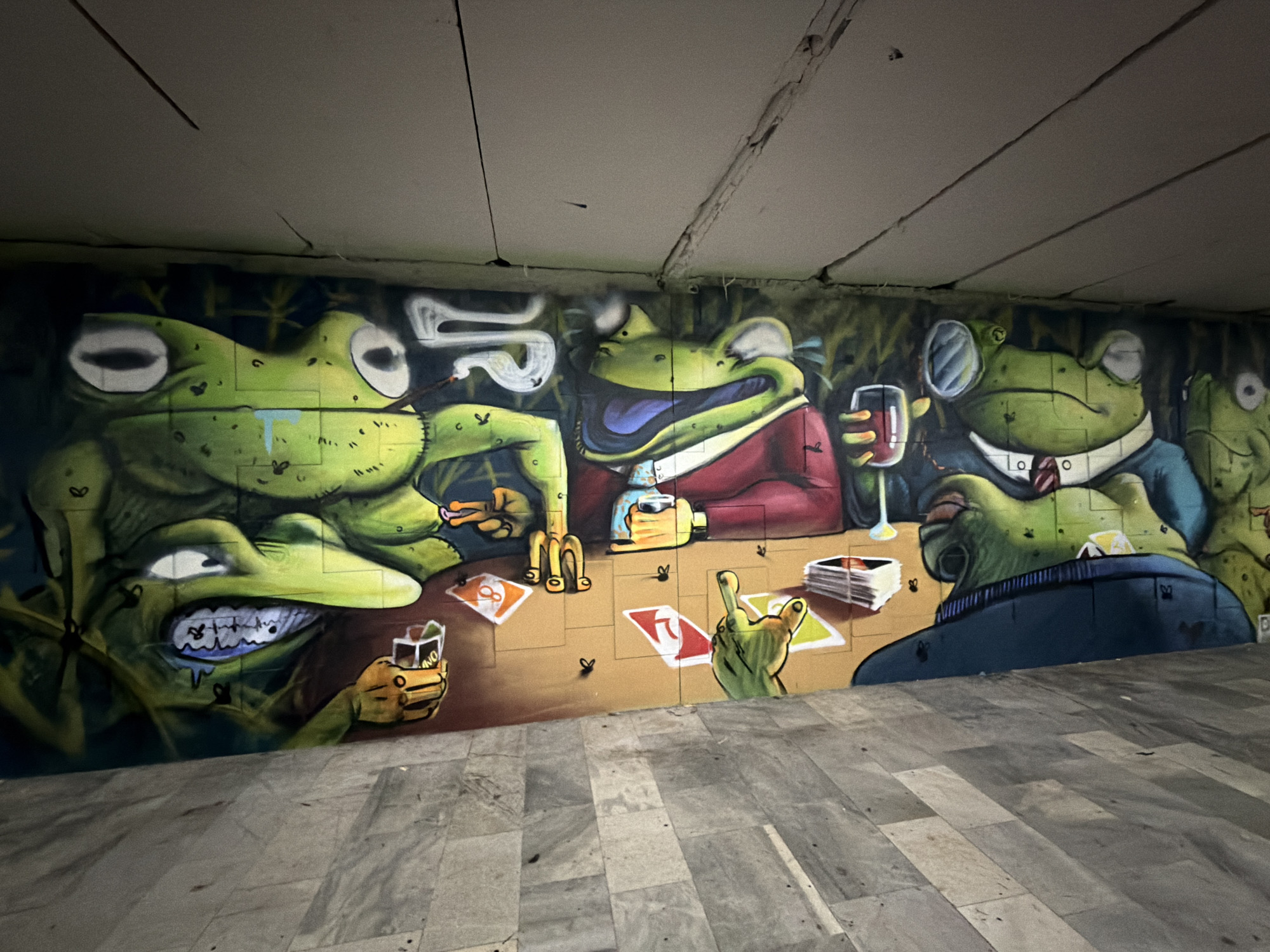 The image captures a graffiti mural depicting anthropomorphic frogs engaged in a card game. The mural is painted on a wall of an underpass in Sofia, Bulgaria, evident from the architectural style.
The mural features five green frogs. The leftmost frog has a prominent underbite, exposing teeth, and holds a glass with the word "UNO" on it. Next to it, another frog is smoking. The central frog, dressed in a red jacket, holds a glass of whiskey and is throwing a card. The frog to the right wears a suit and tie, and a monocle, and is holding a glass of wine. A final frog is partially obscured, visible on the far right. The frogs are seated around a table with playing cards and glasses.
The backdrop is a dark, textured wall, with graffiti elements blending into the background. The floor is tiled with grey rectangular stones.