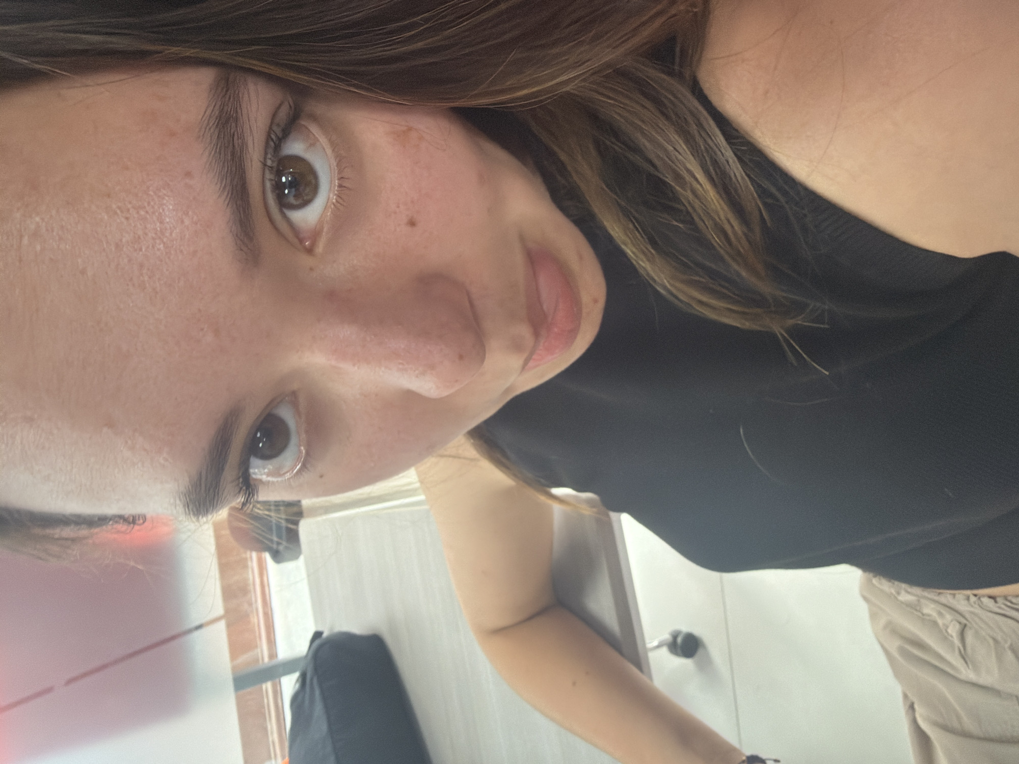 A close-up, angled selfie depicts a woman with fair skin, freckles, and brown hair. She has brown eyes and is wearing a black top. The background is partially visible and includes a black bag, a table with a white surface, and a tiled wall, suggesting an indoor setting. Based on the presence of tiled walls, it is possible that this image was taken in a restaurant or a public space located in the city of San Andrés Cholula, Mexico. However, further details are required to definitively confirm this.