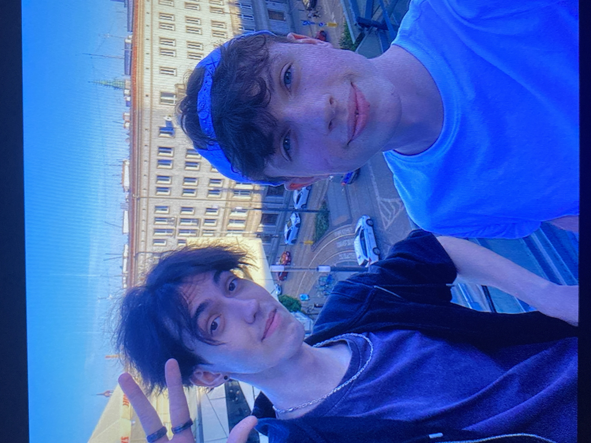 Two young males are taking a selfie on a rooftop overlooking a city street. The male on the left is wearing a dark t-shirt and a black zip-up jacket, with a silver chain necklace. He has dark, messy hair and is making a peace sign with his left hand. The male on the right is wearing a blue t-shirt and a blue bandana on his head. He has light brown, curly hair and is smiling at the camera. The background shows a street with tram tracks, cars, and buildings. The architecture of the buildings, particularly the multi-story, light-colored facade with numerous uniformly spaced windows, suggests a European city. The presence of tram tracks and the general style of the buildings are consistent with the city of Poznan, Poland. The sky is a clear blue, indicating daytime. The scene is captured at an upward angle, looking over the railing of the rooftop.