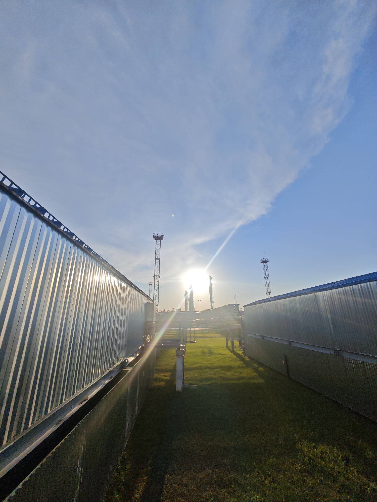 The image shows a scene in Unknown, Russia, featuring industrial structures at sunrise or sunset. The ground is covered in green grass, flanked by two large, corrugated metal buildings on either side. In the distance, between the buildings, are two towers and what appears to be an industrial plant, backlit by the sun. The sky is blue with scattered clouds. A series of pipes and walkways connect the structures in the center. The sun is directly behind the industrial structures, creating a strong lens flare.