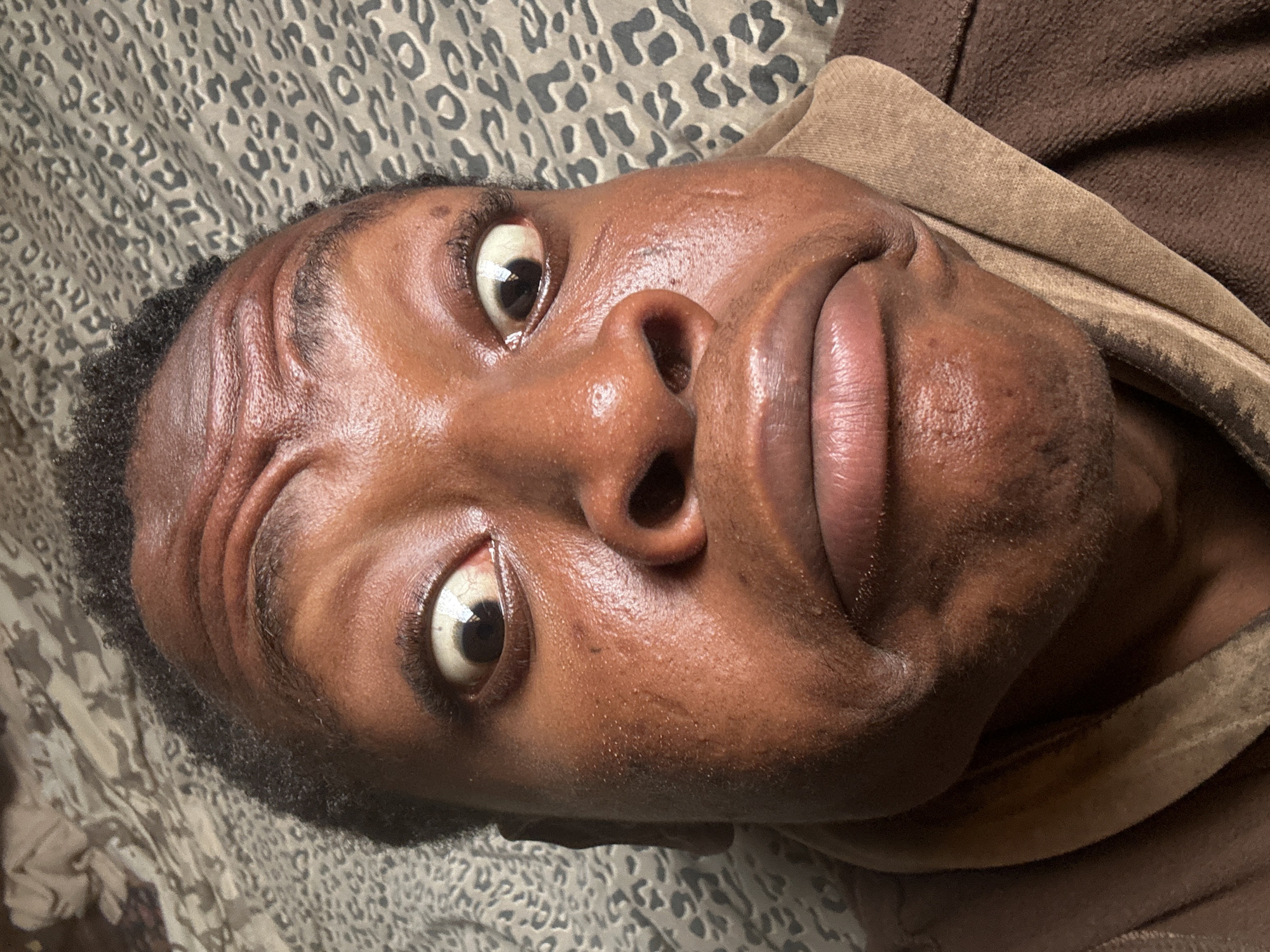 A close-up, high-angle shot shows a Black person's face. The person is lying on a bed with a grey and white patterned bedspread. They are wearing a brown garment with a tan collar. Their eyes are wide and looking upwards. They have a facial expression that is a mix of amusement and curiosity. The subject is indoors and the location is Unknown, Unknown.