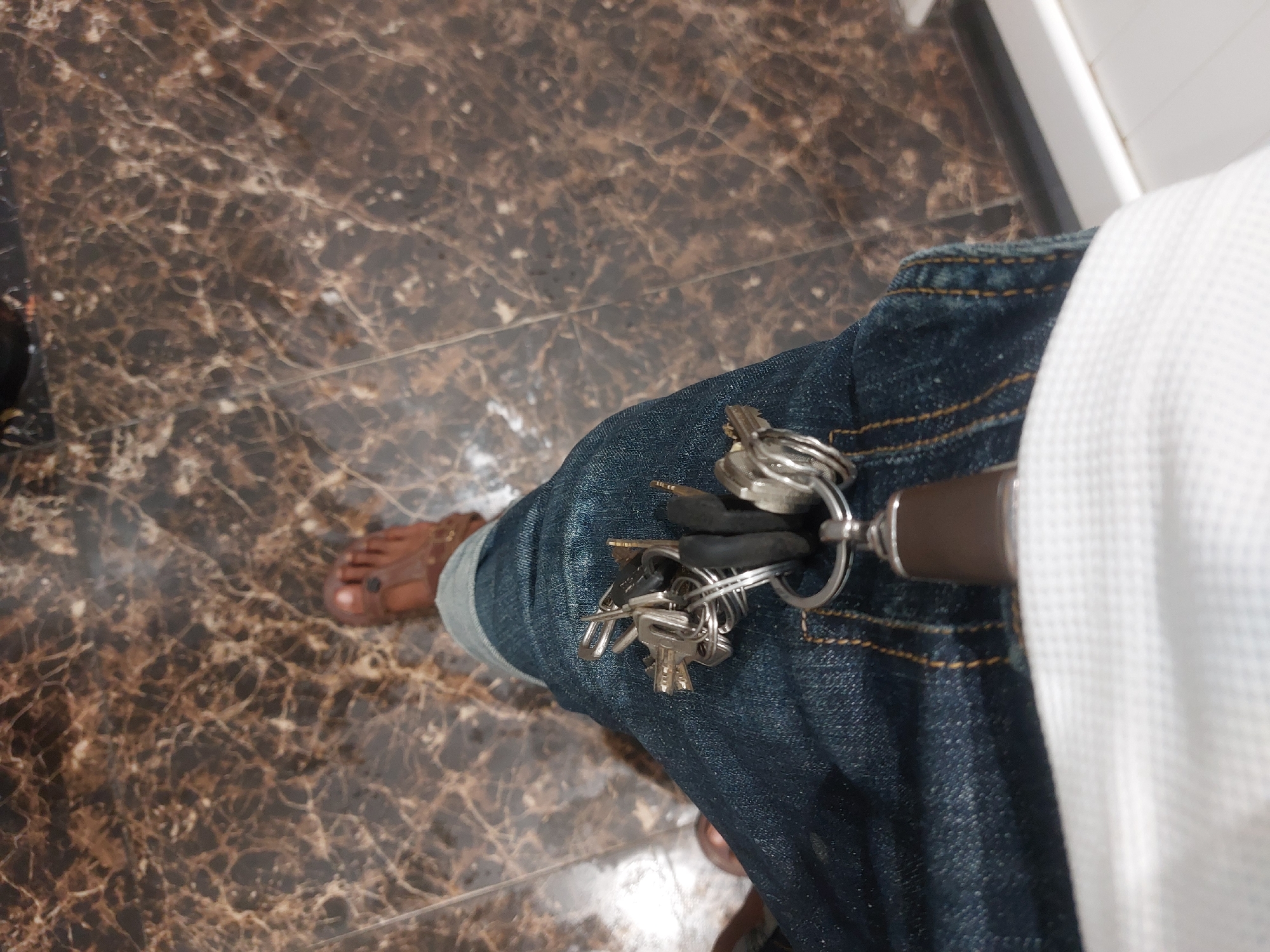 A person wearing jeans and a white shirt is standing on a brown marble-patterned floor. A set of keys hangs from the person's right front pocket. The person is also wearing sandals, exposing their feet. The location cannot be determined from the image, and there is no indication of the city of Dar es-Salaam, Tanzania.
