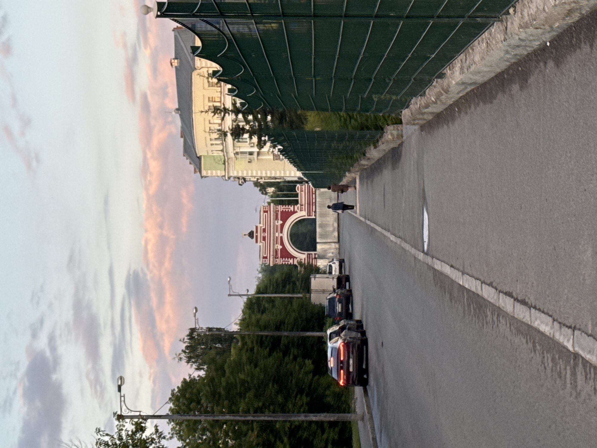 A street scene in Kazan, Russia features a modern road with parked cars and a pedestrian walkway. The sky above displays a soft palette of pink and blue, suggesting either sunrise or sunset. On the left side of the road, several cars are parked along the curb, including a dark-colored SUV and a lighter-colored sedan. Tall, leafy trees line the left edge of the image, and a lamppost stands near the sidewalk.
Further down the street, a large, ornate red and white archway is visible, with intricate detailing. Behind the archway, a building with a light-colored facade and a green roof can be seen.
On the right side of the street, a green metal fence with a decorative top runs along the walkway. Two individuals, one in a dark jacket and pants, and another in a brown outfit, are walking away from the camera on the pedestrian path. A building with a light yellow facade and green accents is situated behind the fence. The overall scene depicts a quiet urban environment with architectural elements and natural scenery.