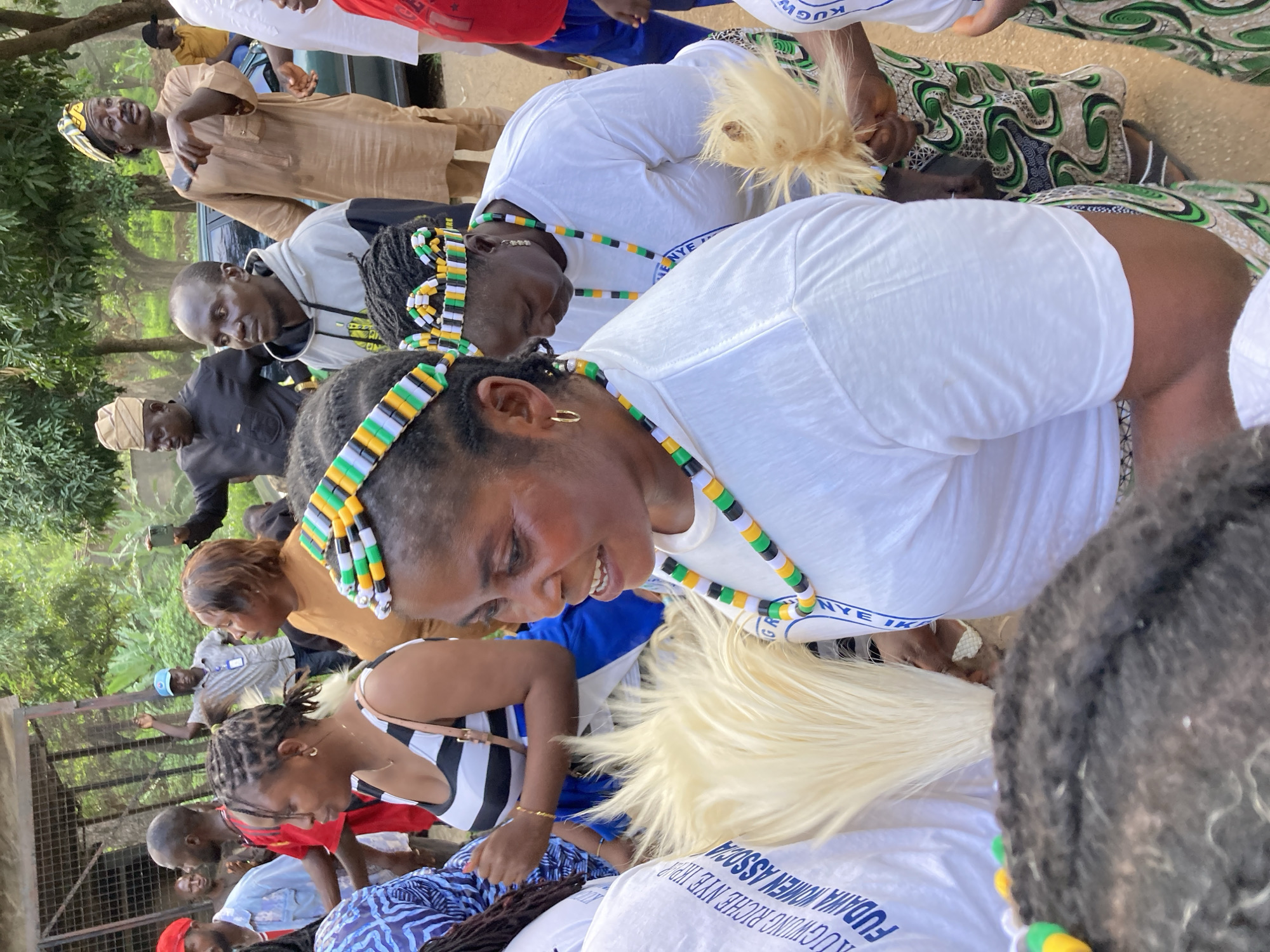 This image depicts an outdoor gathering in what appears to be Jos, Nigeria. The central focus is a group of women adorned with colorful beaded headbands and necklaces, featuring the colors green, yellow, and black. One woman in the foreground, wearing a white t-shirt, has a beaded crown on her head and is holding a whisk-like object made of white hair. Another woman to her left wears a black and white striped top and also sports a beaded headband. In the background, several men are visible, some in traditional attire, including a man in a brown outfit and a conical hat, and another in a black outfit with a similar hat. Some individuals are holding mobile phones, suggesting photography or recording is taking place. The environment appears to be a somewhat informal setting with trees and possibly some structures in the background. The overall atmosphere suggests a cultural event or celebration.