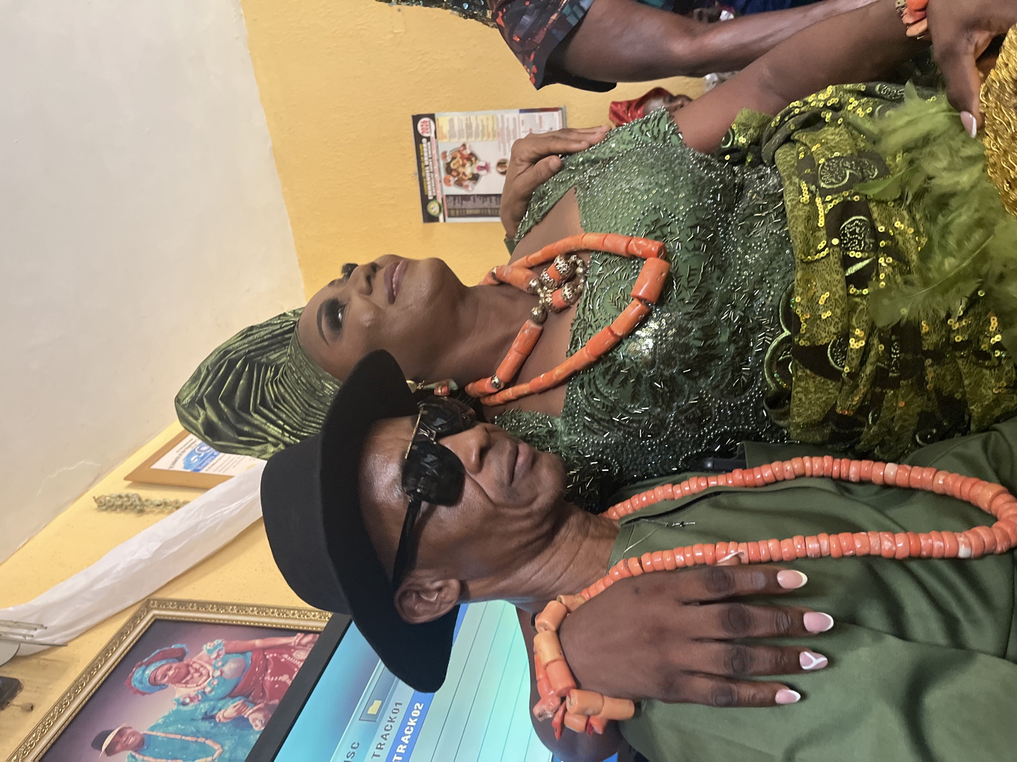 A photograph shows two people, likely a couple, dressed in traditional Nigerian attire, possibly in Warri, Nigeria. A man wears a black fedora hat, sunglasses, an olive-green shirt, and a large coral bead necklace. His manicured hand rests on his chest, with nails painted in a French tip style. Next to him, a woman is adorned in a green, sequined dress, a matching headwrap, and a similar coral bead necklace. The woman looks upward. They are positioned next to a table with framed pictures and a screen showing what appears to be music track selections. A poster or menu is attached to the yellowish wall behind them.