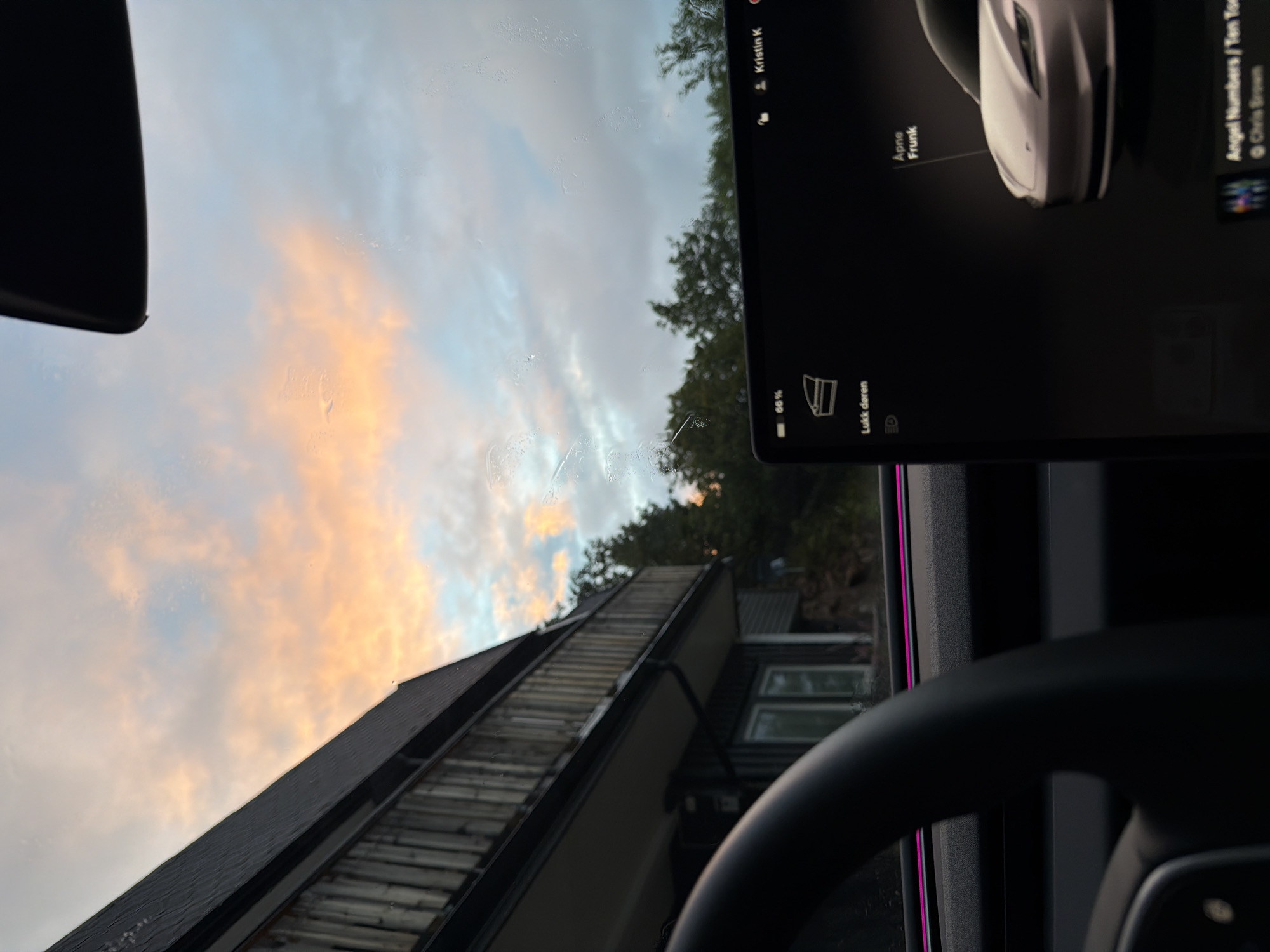 The image shows a view from inside a car, looking out the windshield. The sky is partly cloudy with pastel colors suggesting sunrise or sunset. To the left, a wooden-sided building with a balcony is visible, partially obscured by trees. The car's dashboard is in the foreground, with a large touchscreen display showing a car icon, the words "Apne Frunk," and song information. The location is stated to be in Harstad, Norway.