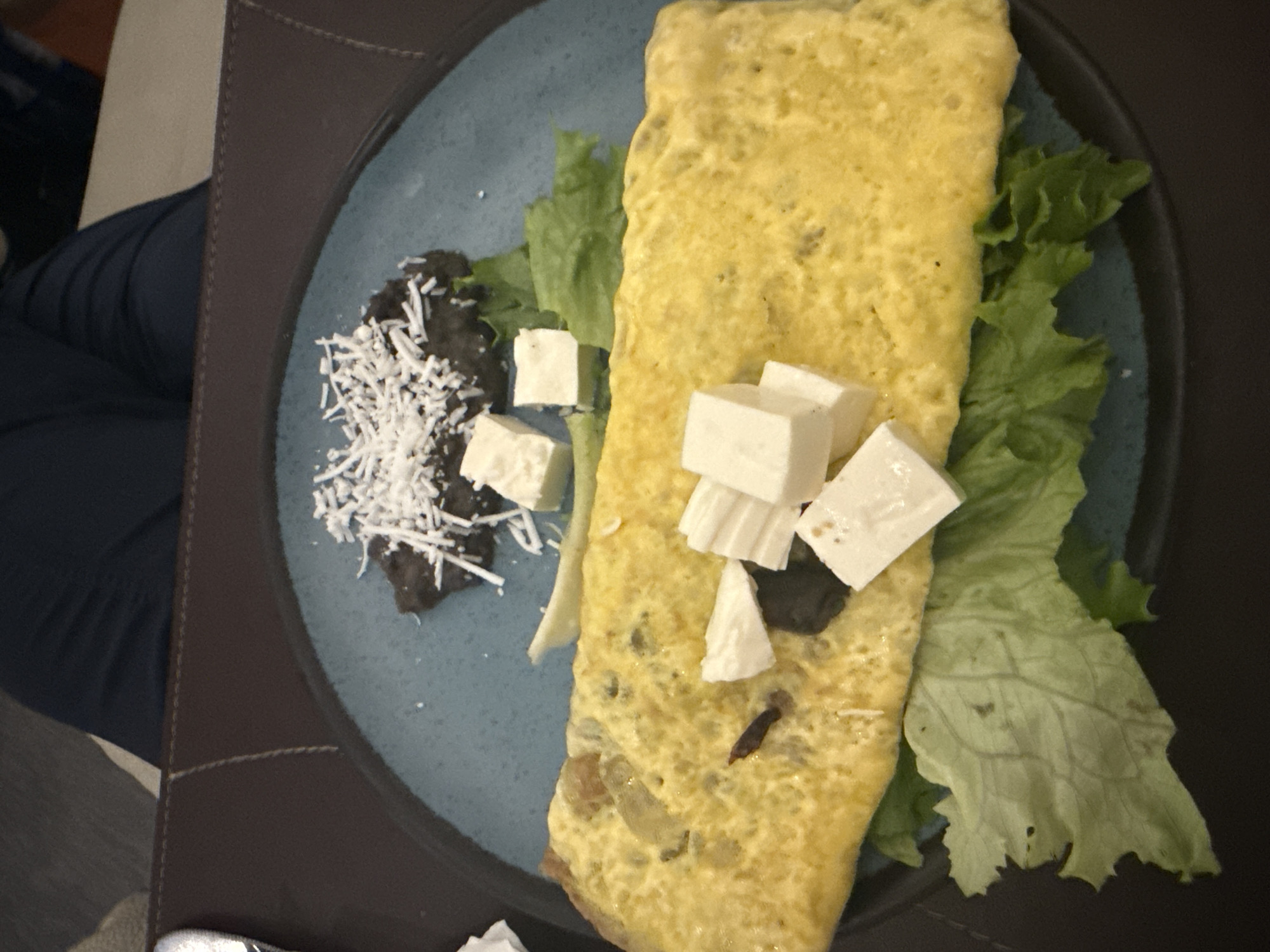 On a blue plate, an omelet is presented alongside black beans topped with shredded cheese, lettuce, and diced white cheese. The plate rests on a dark brown surface. The scene appears to be indoors. There is no indication of this being located in Mexico City.