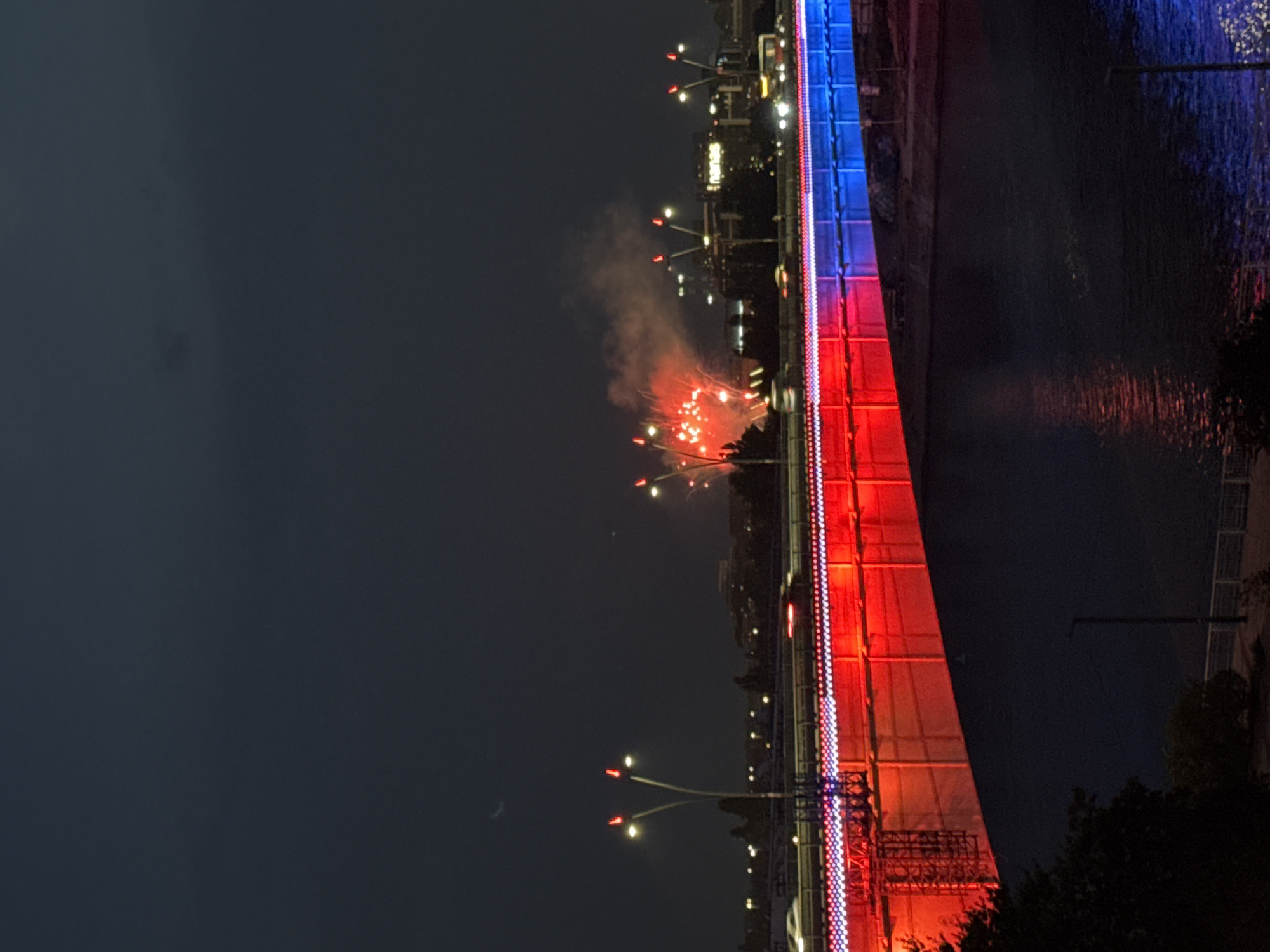 The image shows a nighttime view of the Gazela Bridge in Belgrade, Serbia, adorned with red and blue lights. Fireworks are visible in the sky above the bridge, creating plumes of smoke and bright bursts of light. The bridge itself is illuminated with a gradient of red on the left side transitioning to blue on the right. Traffic lights are visible on the bridge, and cars can be seen moving along it, their headlights and taillights creating streaks of light. The dark sky above is dotted with a few faint stars and a sliver of the moon. The river beneath the bridge reflects the colorful lights. The urban landscape of Belgrade is visible in the background, with illuminated buildings and streetlights. The scene suggests a festive occasion, likely a celebration involving fireworks.