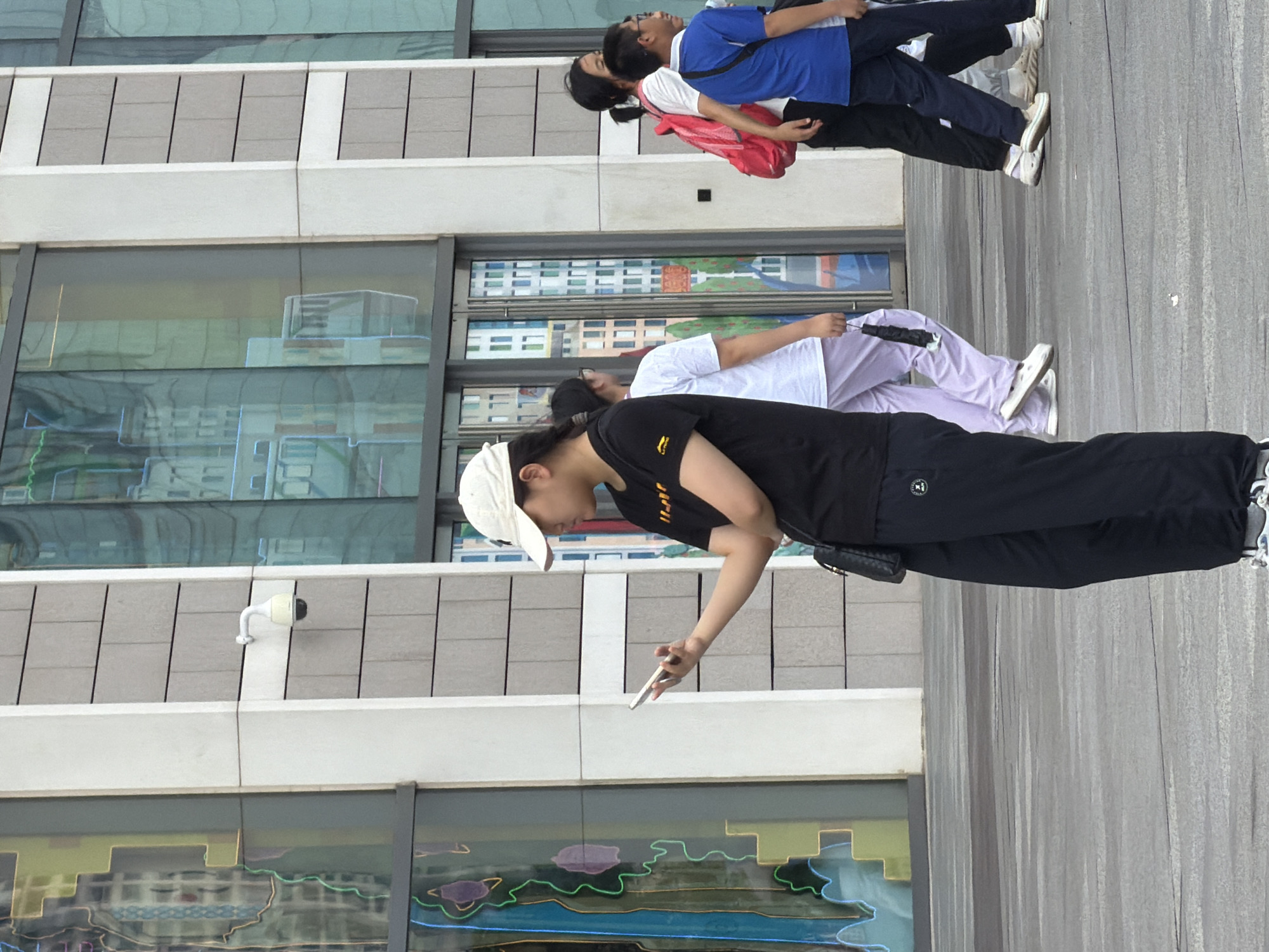 A group of people walk across a grey, paved public square in front of a modern building in Yuzhong District, China. The building features large glass windows with colorful artistic patterns resembling buildings. A young woman in a black t-shirt and dark pants stands in the foreground, looking at her smartphone. To her right, a person in a white t-shirt and light purple pants walks away from the camera, holding a black item. Further to the right, two young men walk towards the camera, one in a white t-shirt and a pink backpack, and the other in a blue t-shirt and dark pants. A security camera is mounted on the building wall to the left of the group. The lighting suggests it is daytime.