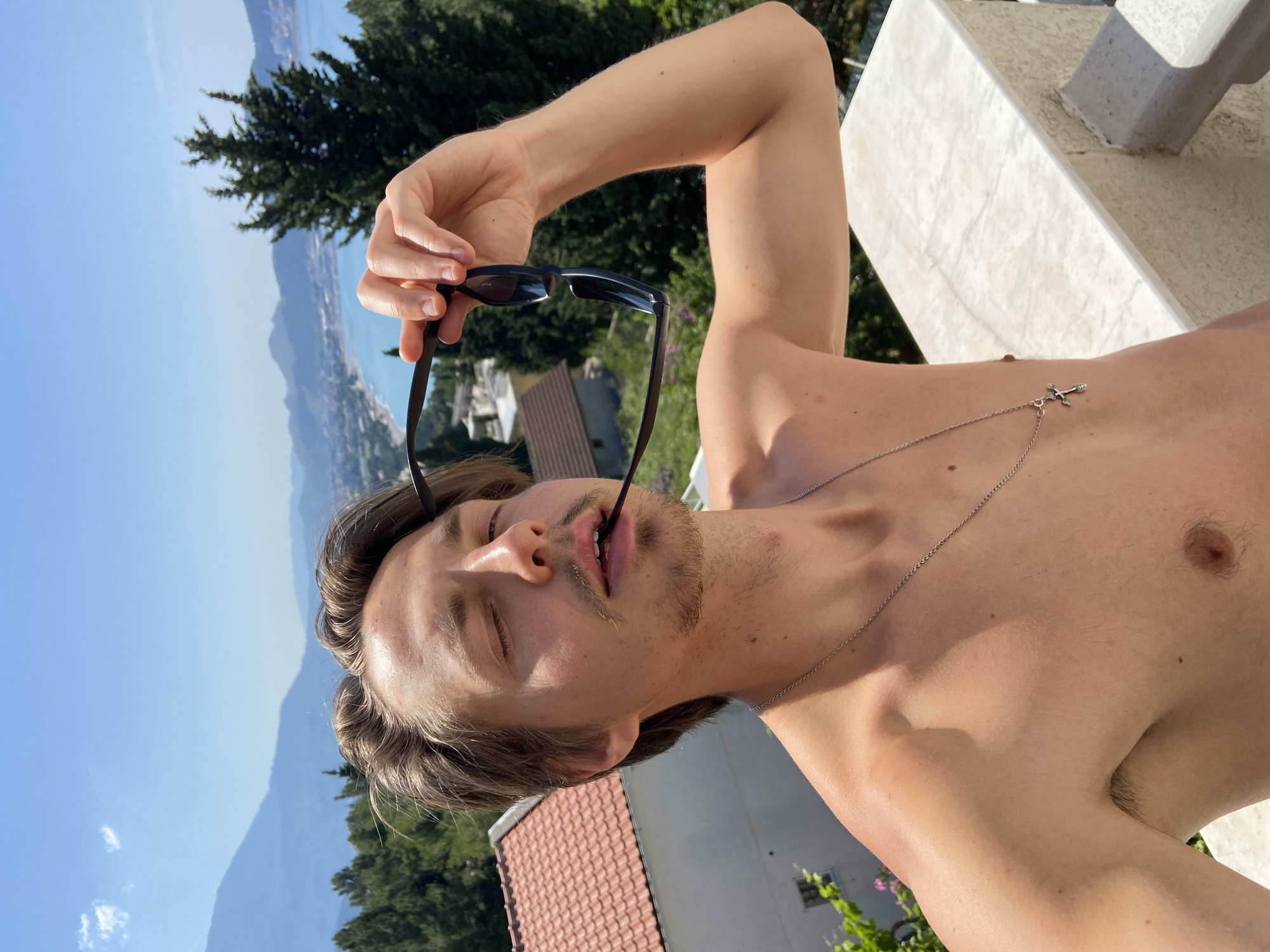 A topless male with fair skin, light brown hair, and a light beard holds sunglasses in his mouth. He wears a silver necklace with a cross pendant. The backdrop includes a mountain range, trees, building rooftops, and a section of the Adriatic Sea. The picture appears to be taken in a sunny outdoor setting. It is not possible to confirm that the exact location is Šušanj, Montenegro from the image alone.